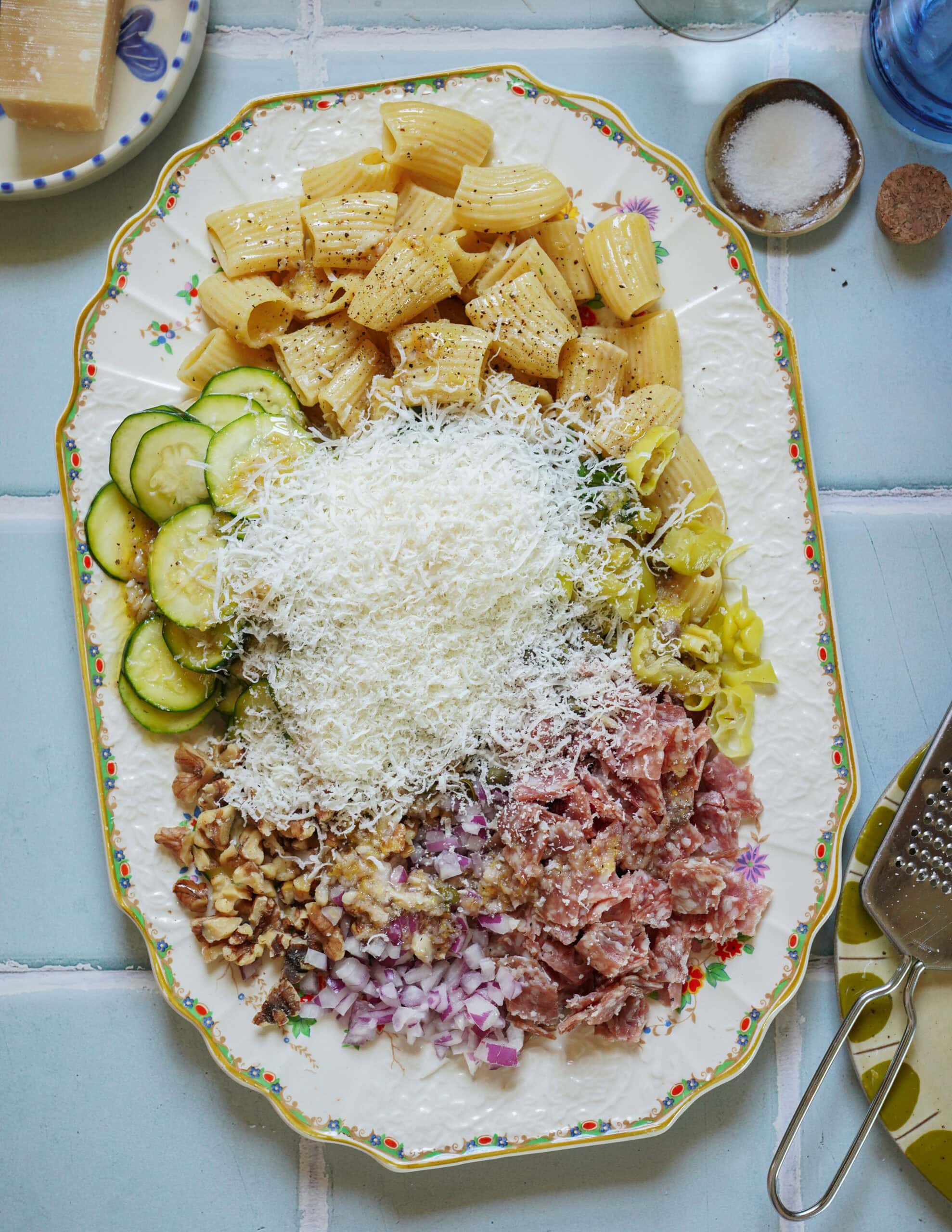 A decorative plate holds rigatoni pasta, sliced zucchini, chopped walnuts, diced red onions, crumbled sausage, pepperoncini, and a large mound of grated cheese, arranged in separate sections on a light blue tiled table.