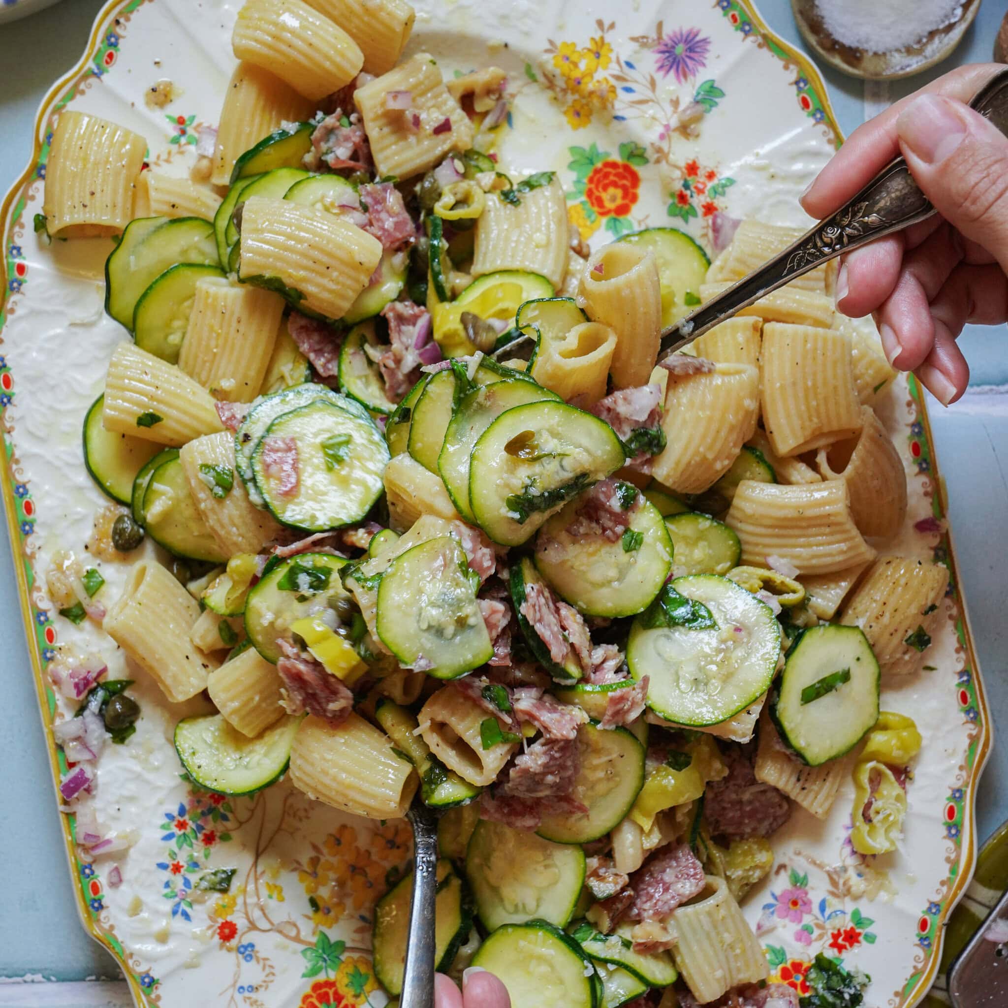 A floral plate filled with pasta, sliced zucchini, salami, capers, and herbs. Two hands are serving the pasta with forks from opposite sides of the plate.