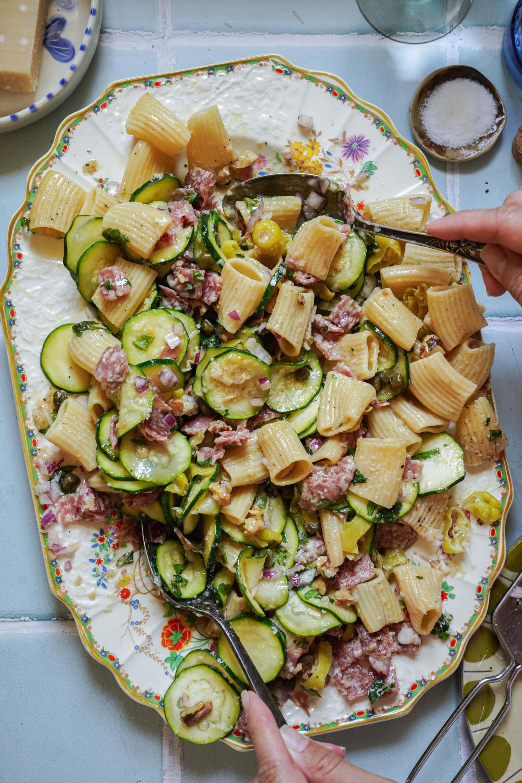 A floral plate holds pasta with rigatoni, zucchini slices, red onions, cured meat, green olives, and herbs. Two hands serve the salad using silver utensils. Salt and cheese are visible nearby on a light blue tiled surface.