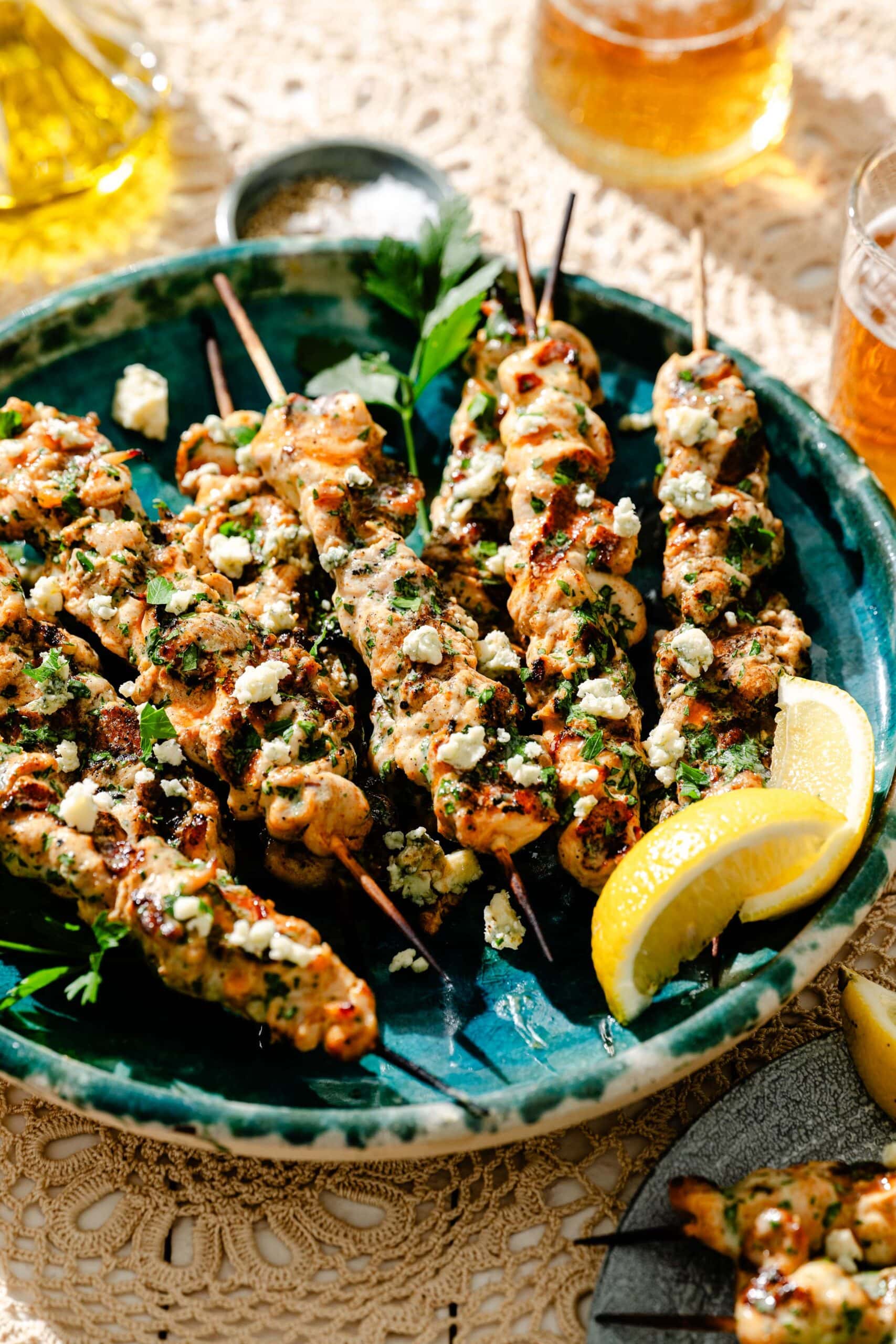 Grilled chicken skewers topped with herbs and crumbled cheese are served on a green plate with lemon wedges. Drinks and seasonings are visible in the background on a lace tablecloth.