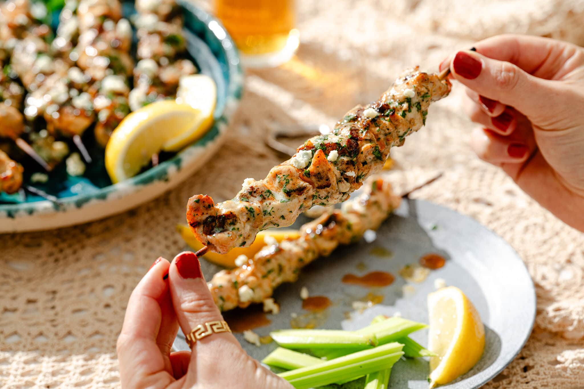 A person holds a grilled chicken skewer over a plate with celery sticks and a lemon wedge; more skewers and a glass of beer are in the background.