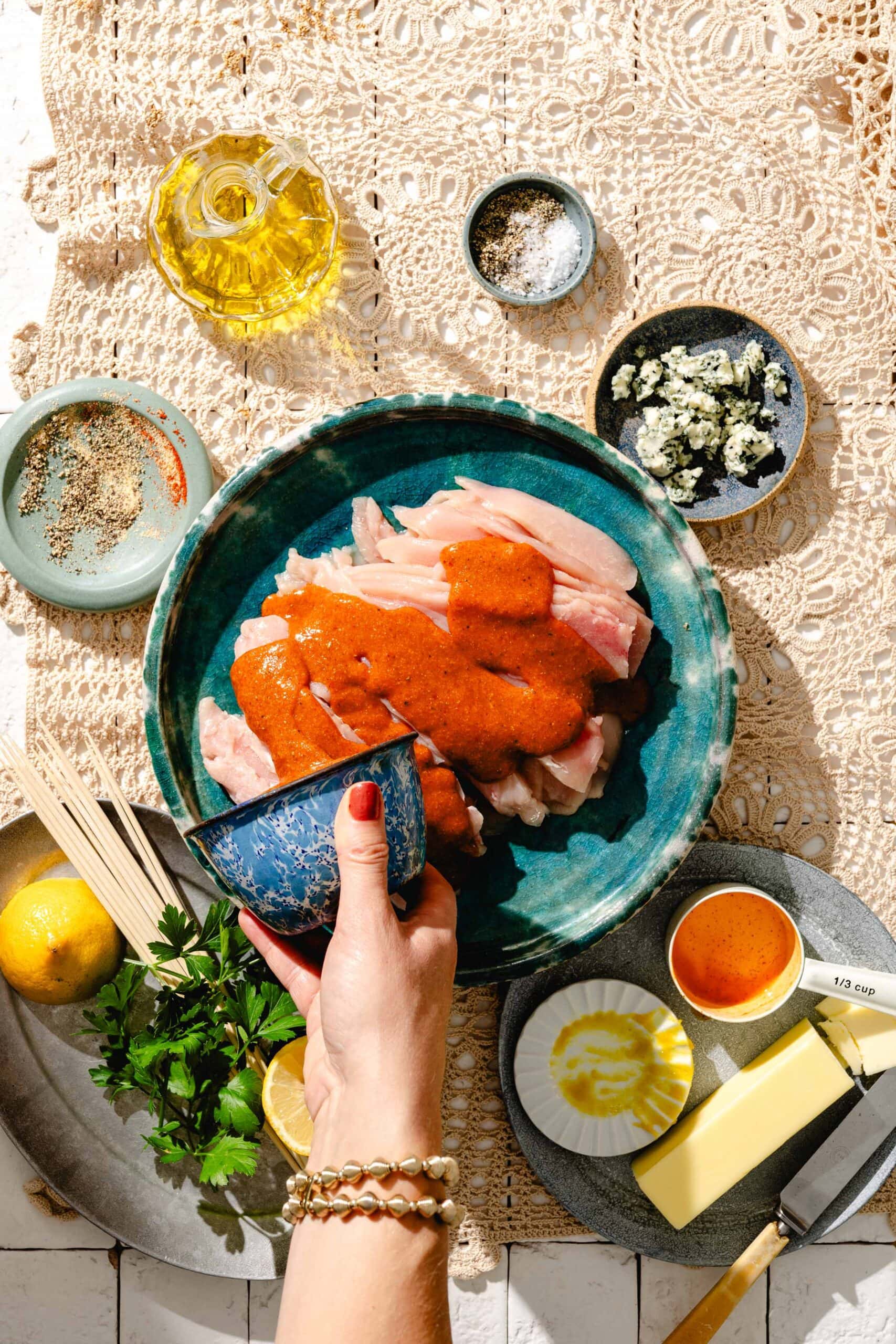A hand pours sauce over raw chicken in a teal bowl, surrounded by butter, herbs, lemon, olive oil, blue cheese, spices, and a knife on a lace tablecloth. Preparing ingredients for a meal.