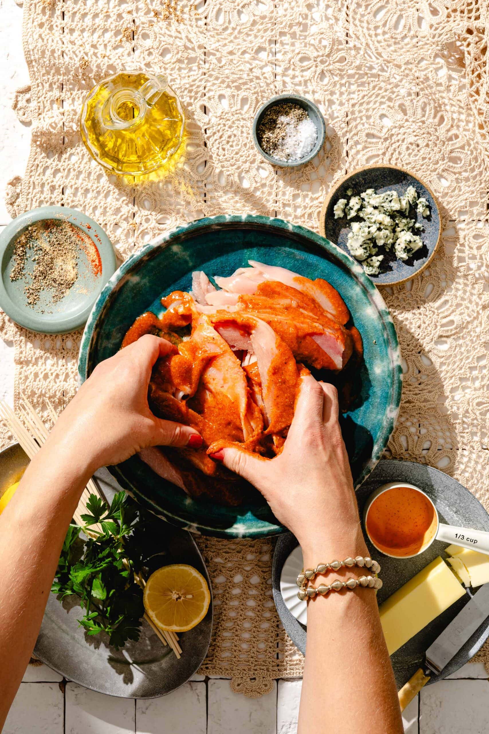 Hands mixing seasoned raw chicken in a blue bowl, surrounded by small dishes of spices, butter, herbs, lemon, blue cheese, and oil on a lacy beige tablecloth.