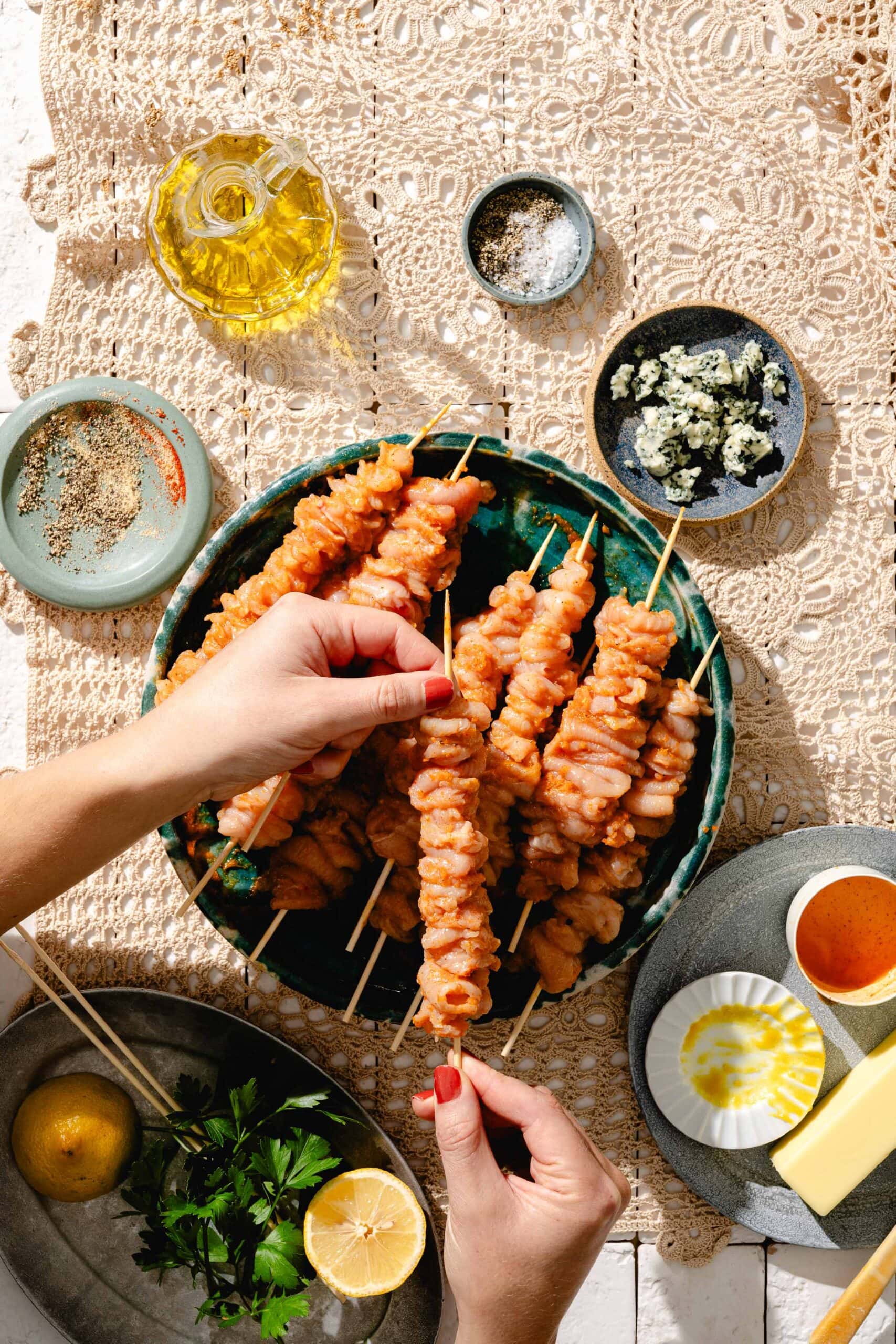 Hands arranging cooked chicken skewers on a green plate, surrounded by bowls of spices, blue cheese, herbs, lemon halves, butter, honey, and olive oil on a lace tablecloth.