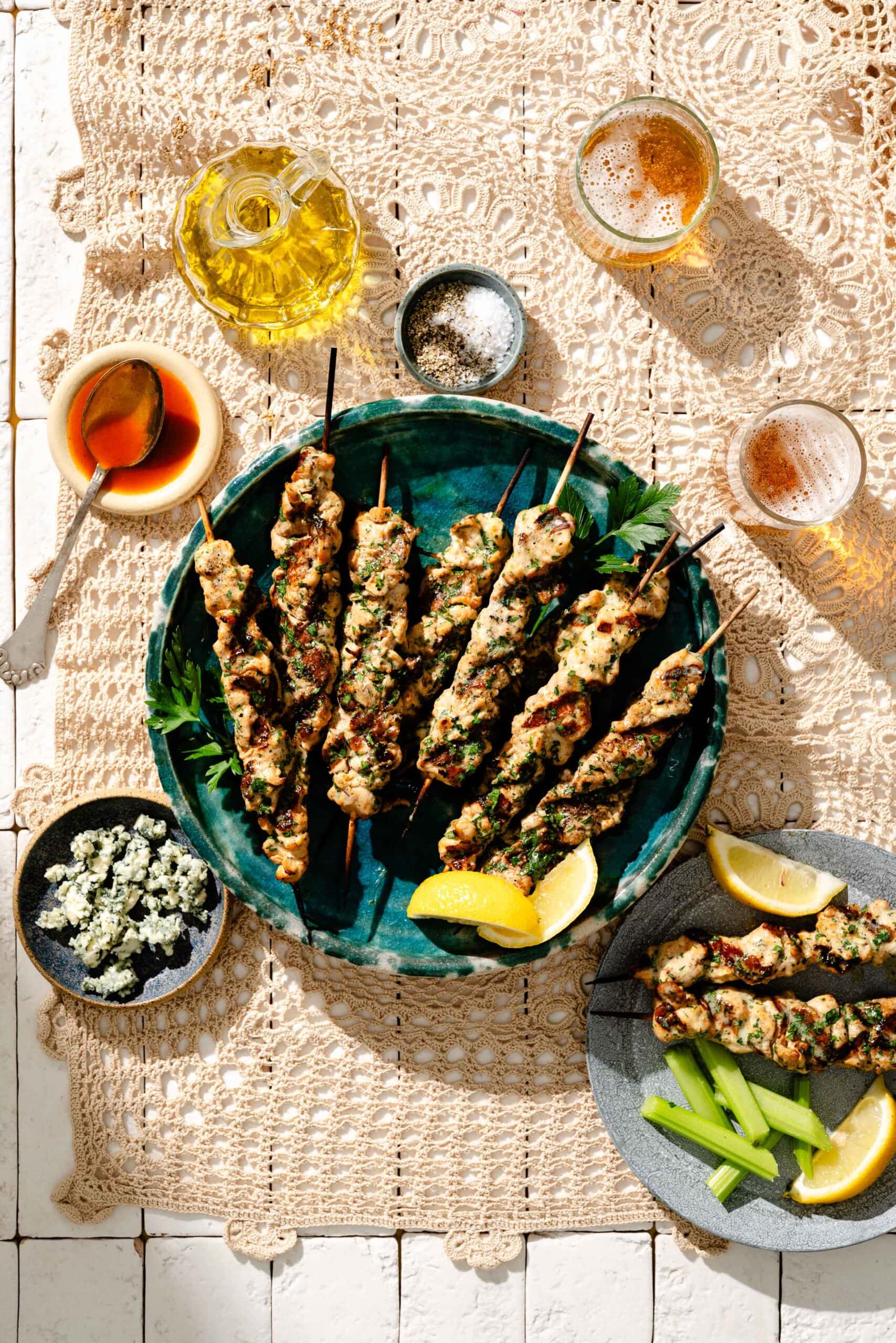 Grilled chicken skewers on a green plate with lemon wedges, surrounded by celery sticks, blue cheese, oil, honey, salt, and drinks on a lace tablecloth.