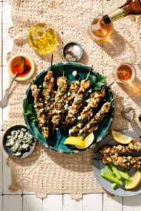A plate of grilled chicken skewers topped with herbs and cheese, served with lemon wedges, celery sticks, dipping sauces, and drinks on a lace tablecloth.