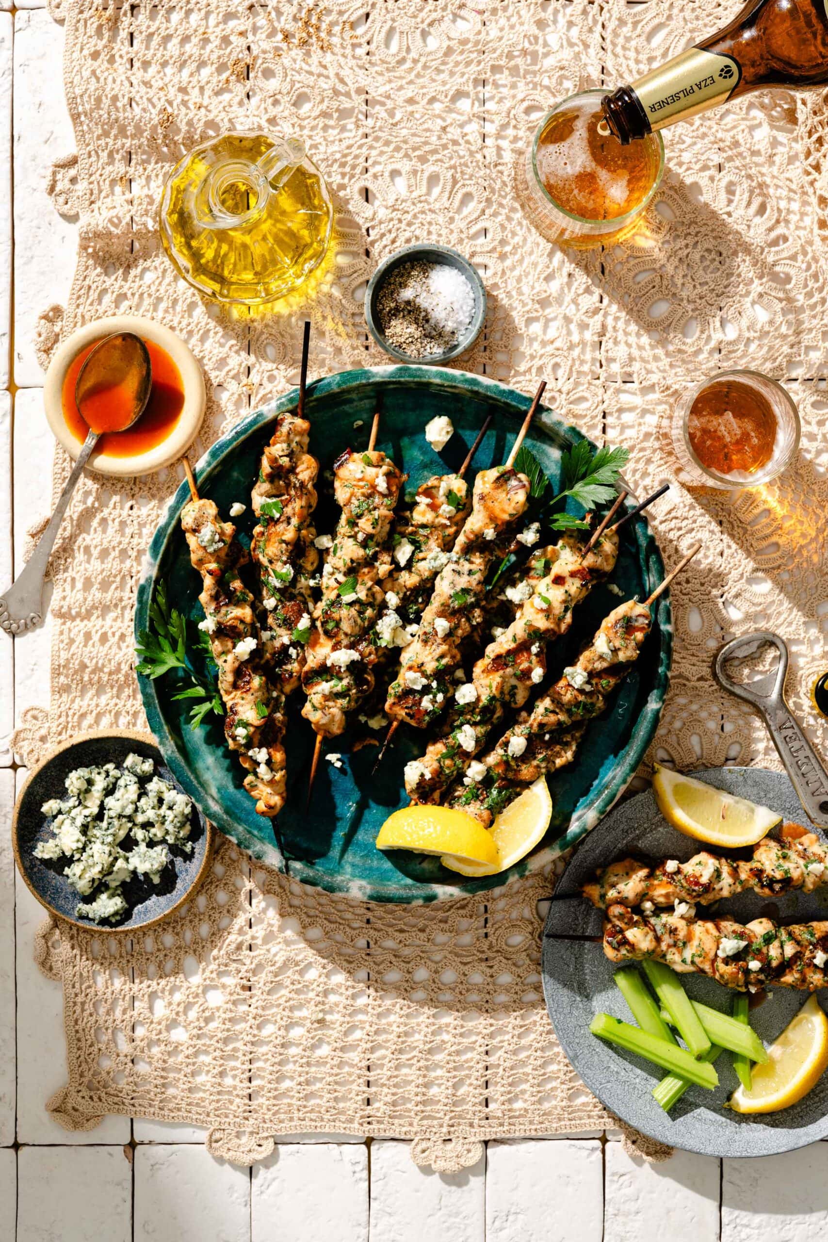 A plate of grilled chicken skewers topped with herbs and cheese, served with lemon wedges, celery sticks, dipping sauces, and drinks on a lace tablecloth.