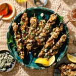 Grilled chicken skewers topped with herbs and cheese are arranged on a green plate with lemon wedges, surrounded by small dishes of blue cheese, sauce, and spices on a lace tablecloth.