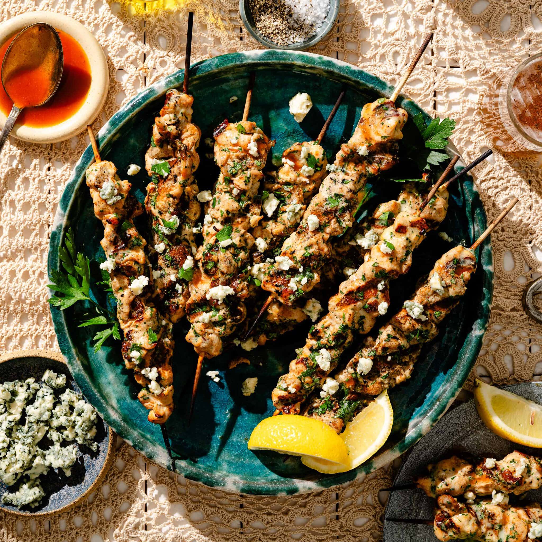 Grilled chicken skewers topped with herbs and cheese are arranged on a green plate with lemon wedges, surrounded by small dishes of blue cheese, sauce, and spices on a lace tablecloth.