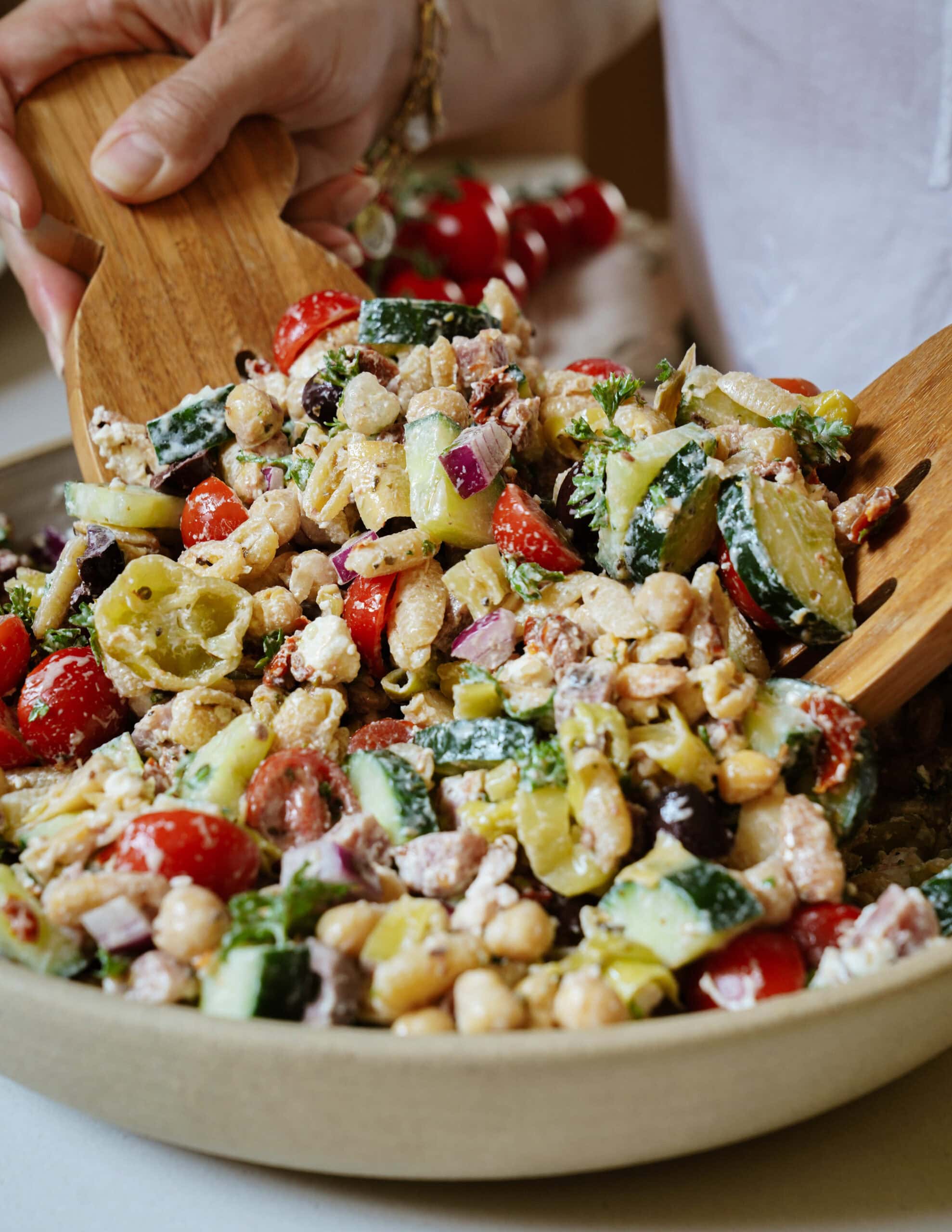 A person tosses a colorful chickpea salad with tomatoes, cucumbers, red onion, olives, feta, and pepperoncini in a large bowl using wooden salad servers.