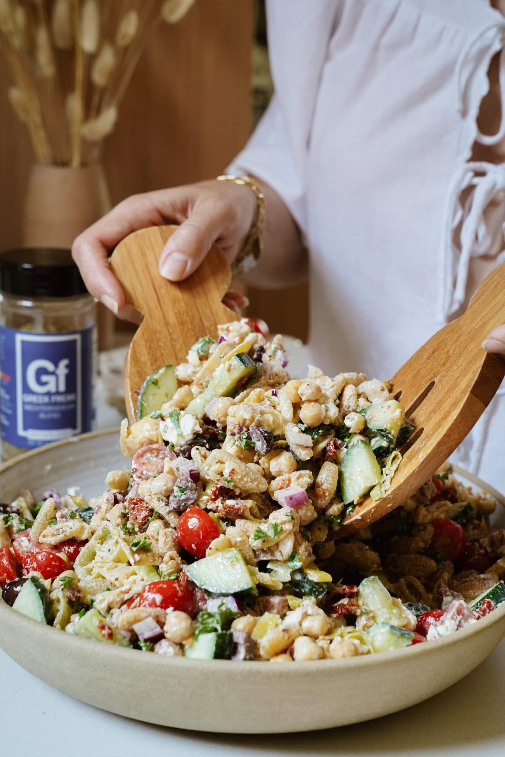 A person in a white shirt tosses a colorful chickpea salad with wooden utensils. A jar labeled Gf sits nearby on the table. The salad contains cucumbers, tomatoes, chickpeas, and red onions.