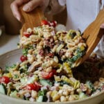 A person serves a colorful chickpea salad with cherry tomatoes, cucumbers, peppers, olives, and herbs from a bowl using wooden salad tongs.