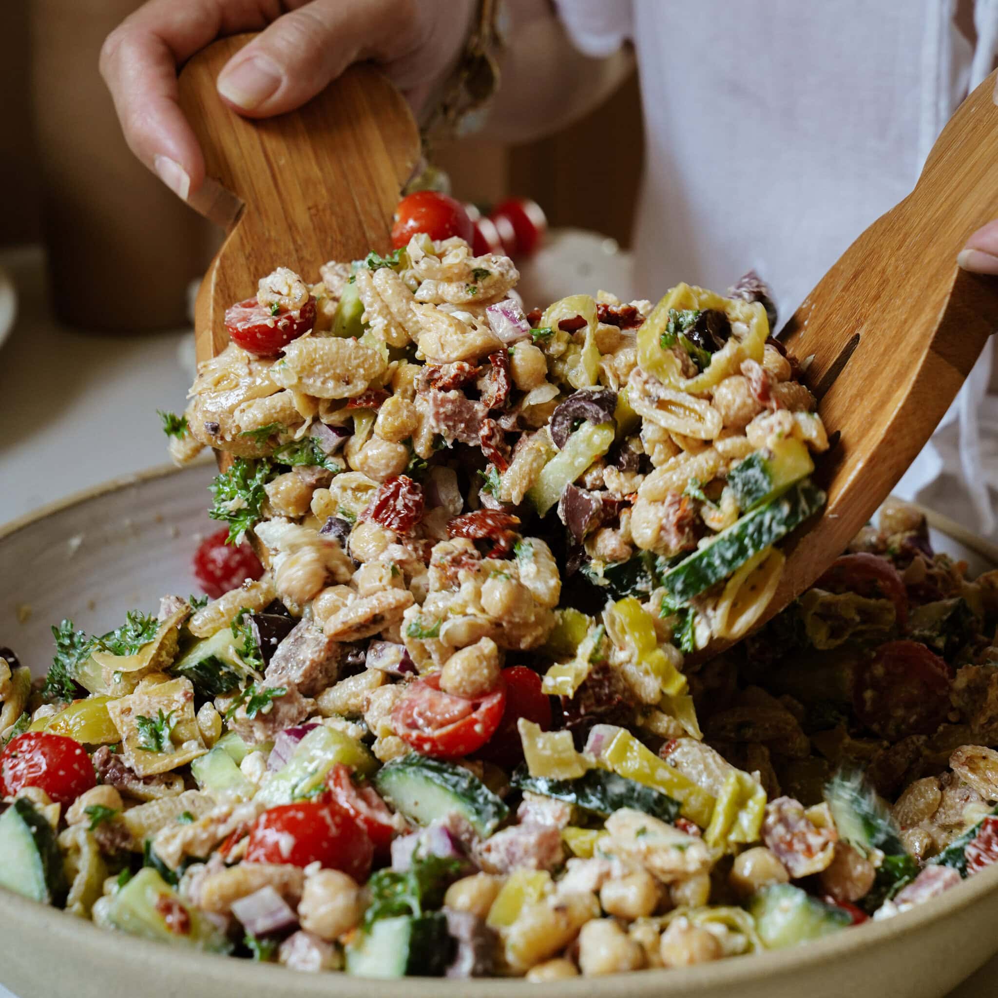 A person serves a colorful chickpea salad with cherry tomatoes, cucumbers, peppers, olives, and herbs from a bowl using wooden salad tongs.