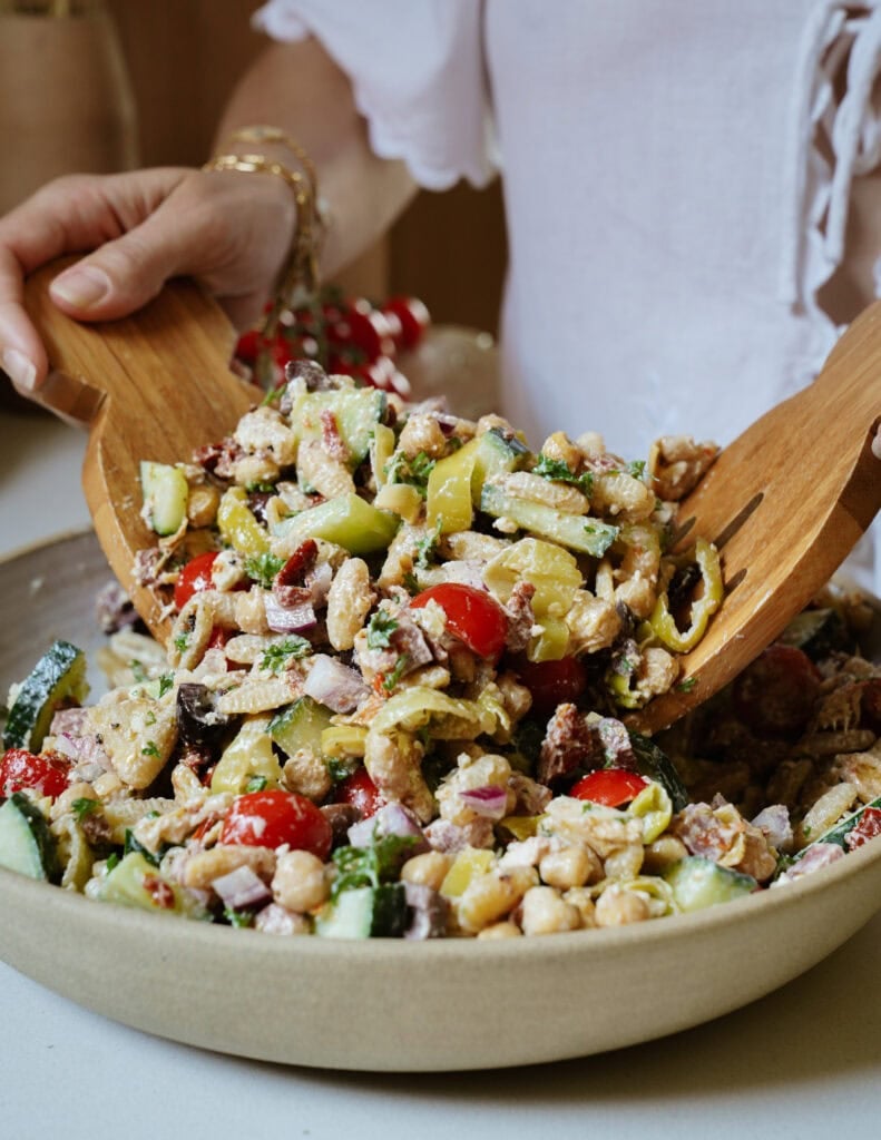 A person in a white shirt tosses a colorful chickpea salad with wooden salad servers. The salad includes cherry tomatoes, cucumber, red onion, and green peppers in a beige bowl.