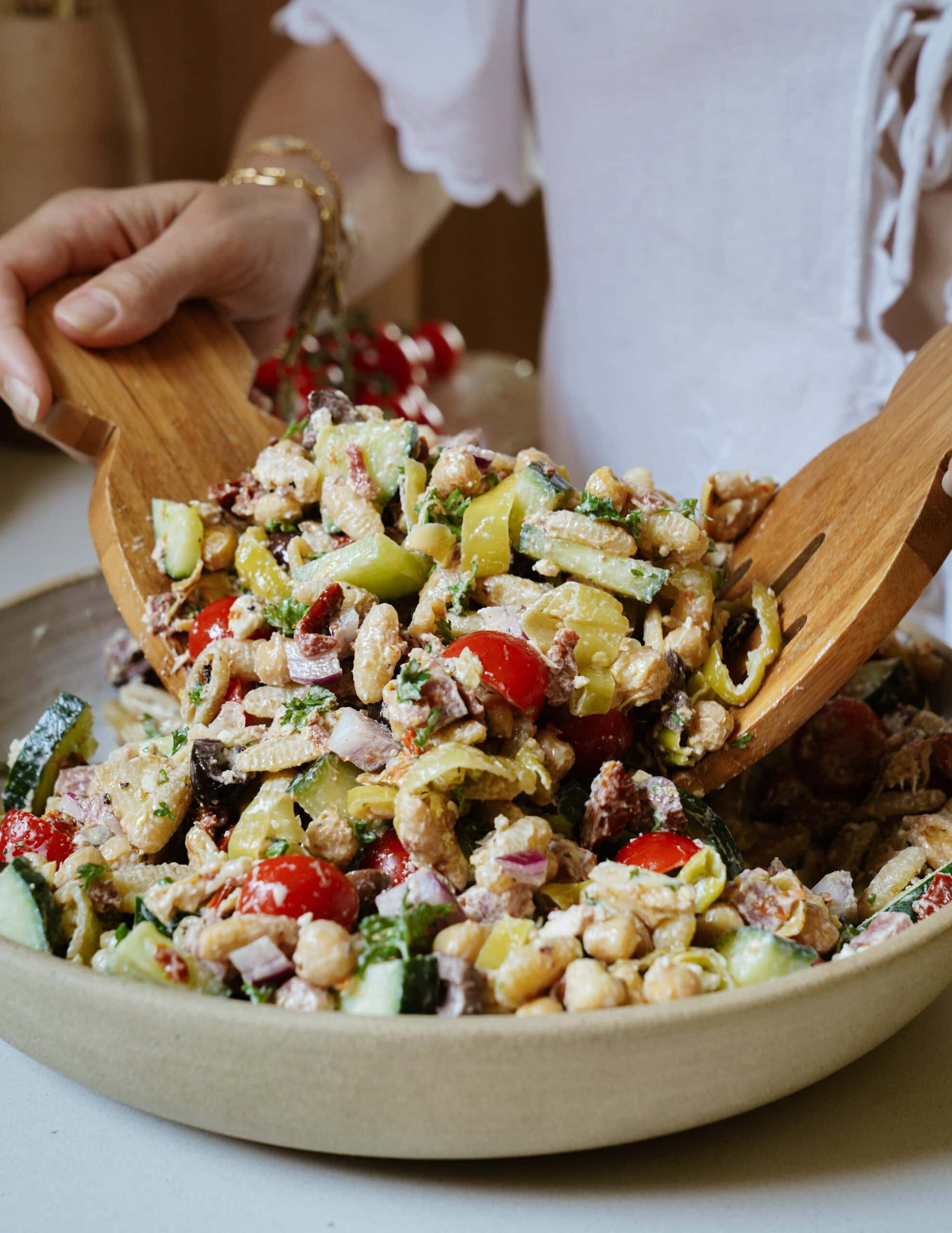 A person in a white shirt tosses a colorful chickpea salad with wooden salad servers. The salad includes cherry tomatoes, cucumber, red onion, and green peppers in a beige bowl.