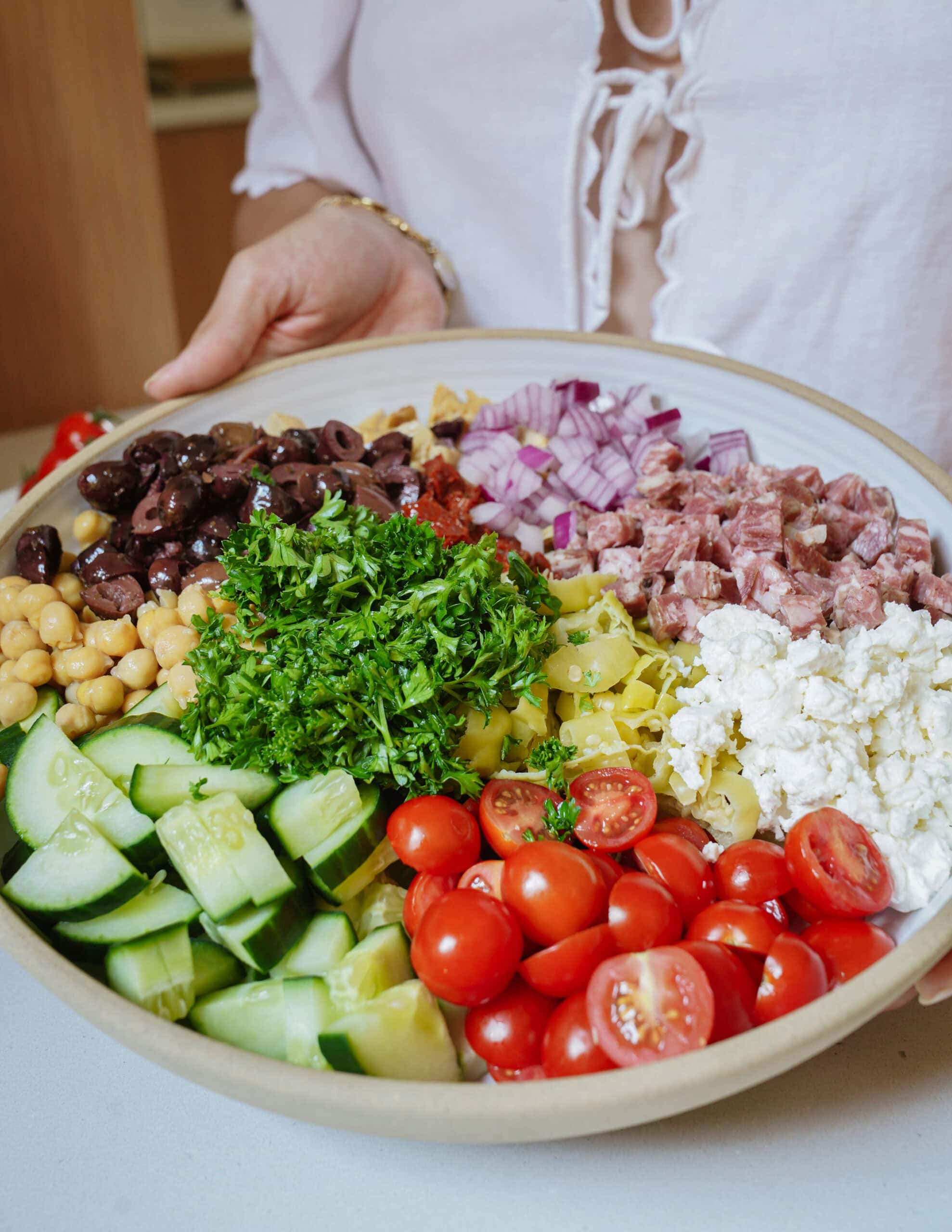 A bowl with cucumber, cherry tomatoes, feta, chickpeas, red onion, olives, salami, parsley, and pepperoncini. A person in a white shirt holds the bowl.