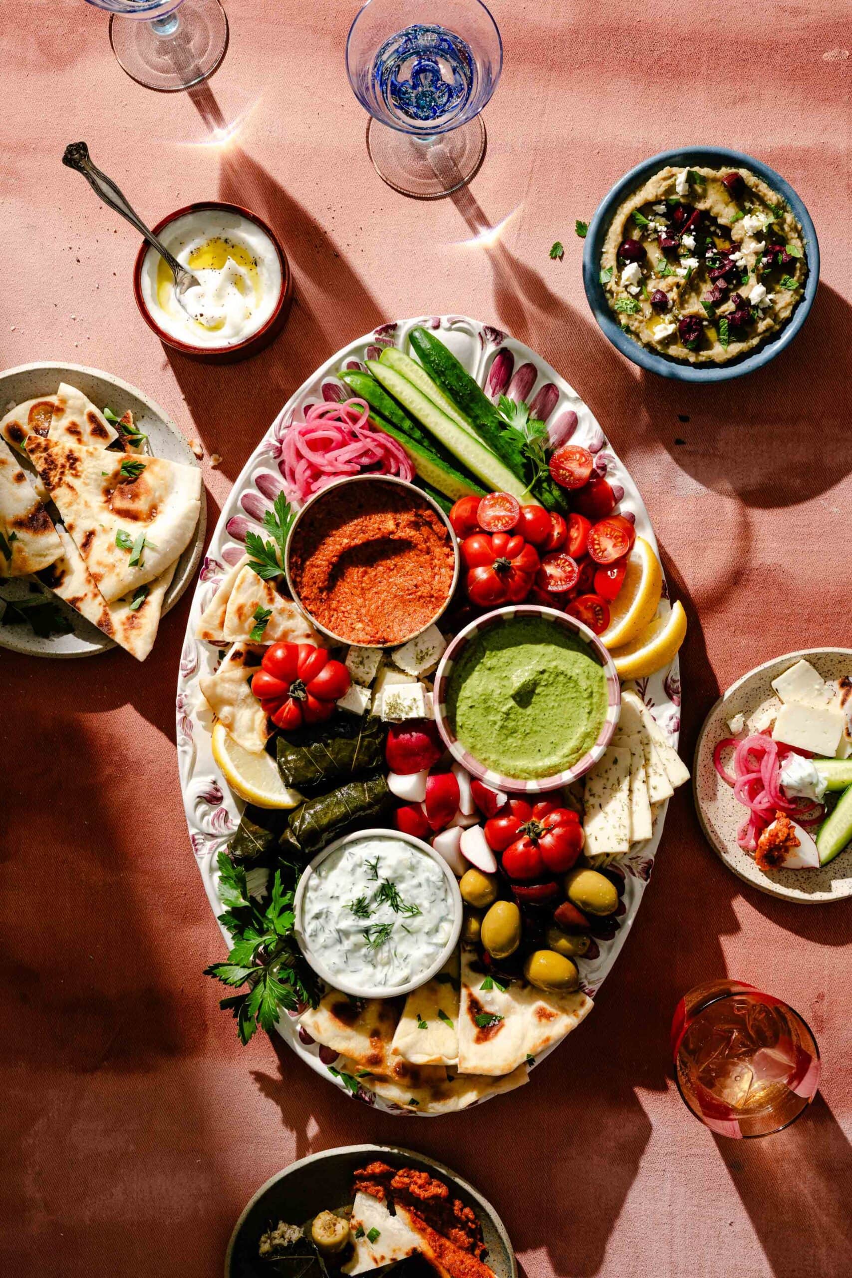 A platter with pita, dips, grape leaves, vegetables, olives, tomatoes, cheese, and herbs is surrounded by plates of hummus, dips, and flatbread on a pink table. Glasses with drinks are also visible.