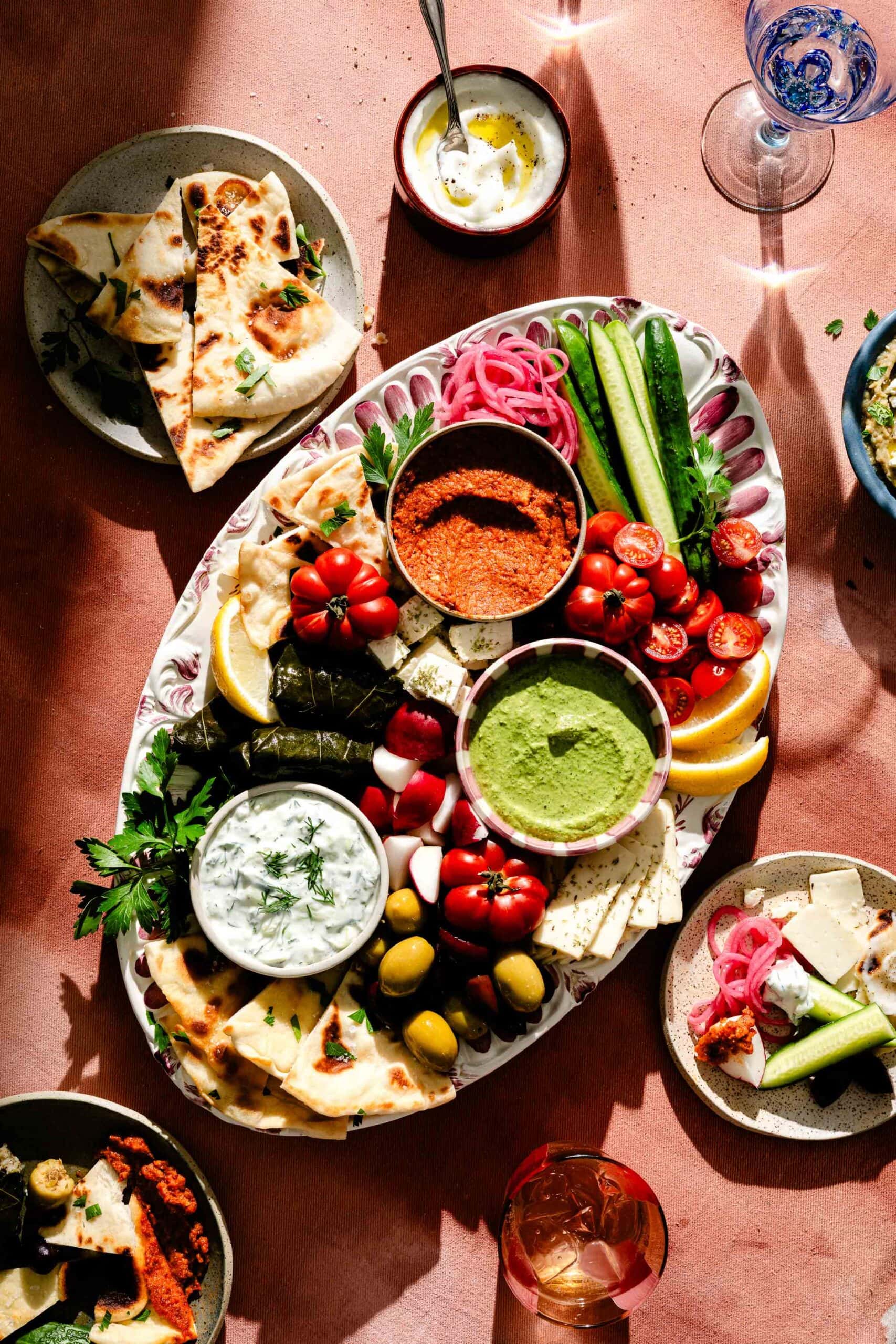 A platter with pita, dips, grape leaves, cucumbers, tomatoes, olives, feta, and herbs, surrounded by small dishes of dips and bread slices on a sunlit table.