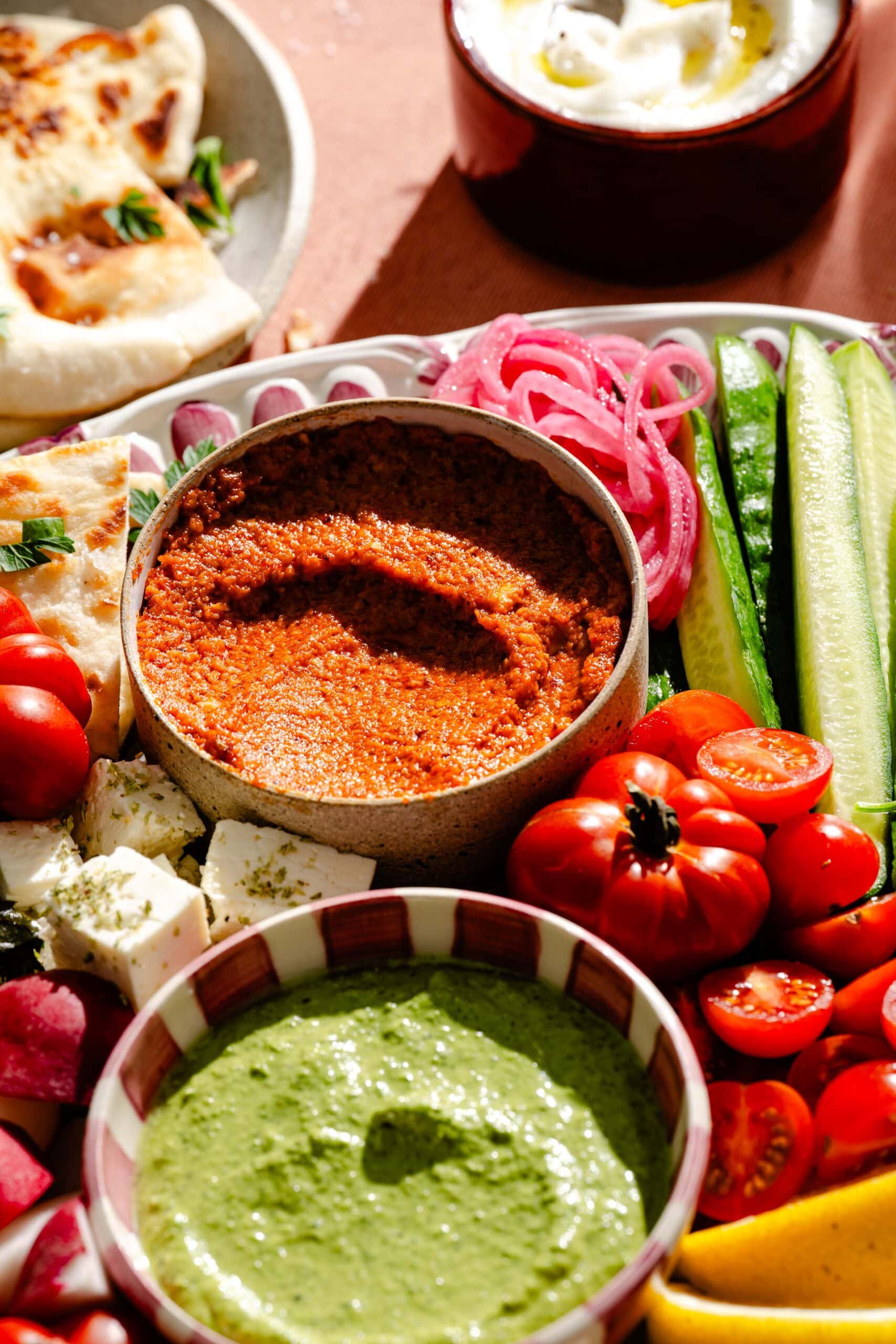 A colorful spread of Mediterranean foods, including green dip, red spread, cucumber sticks, cherry tomatoes, cubed feta cheese, pickled onions, flatbread, and a bowl of yogurt.