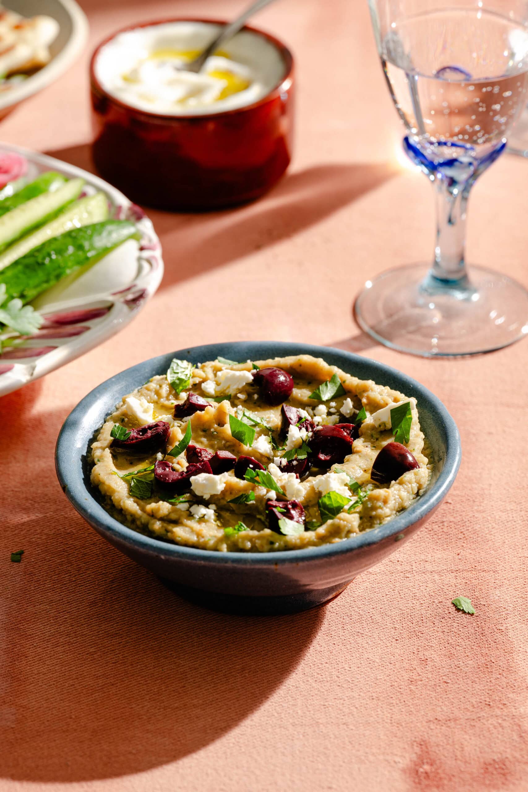 A bowl of hummus topped with olives, feta, and herbs sits on a peach-colored surface near a glass of water and plates of vegetables and yogurt.