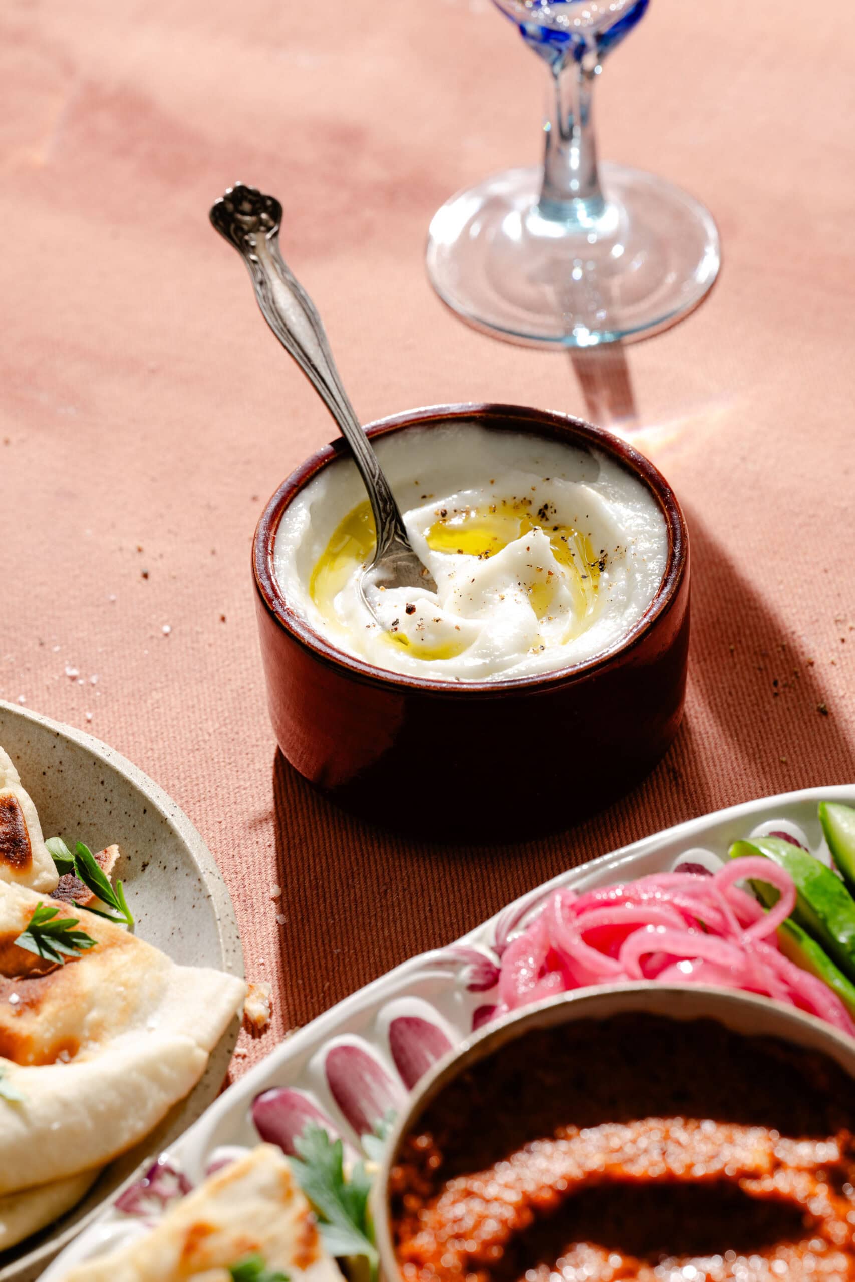 A bowl of yogurt topped with olive oil and pepper sits on a table next to a glass, with plates of naan, pickled onions, and vegetables nearby.