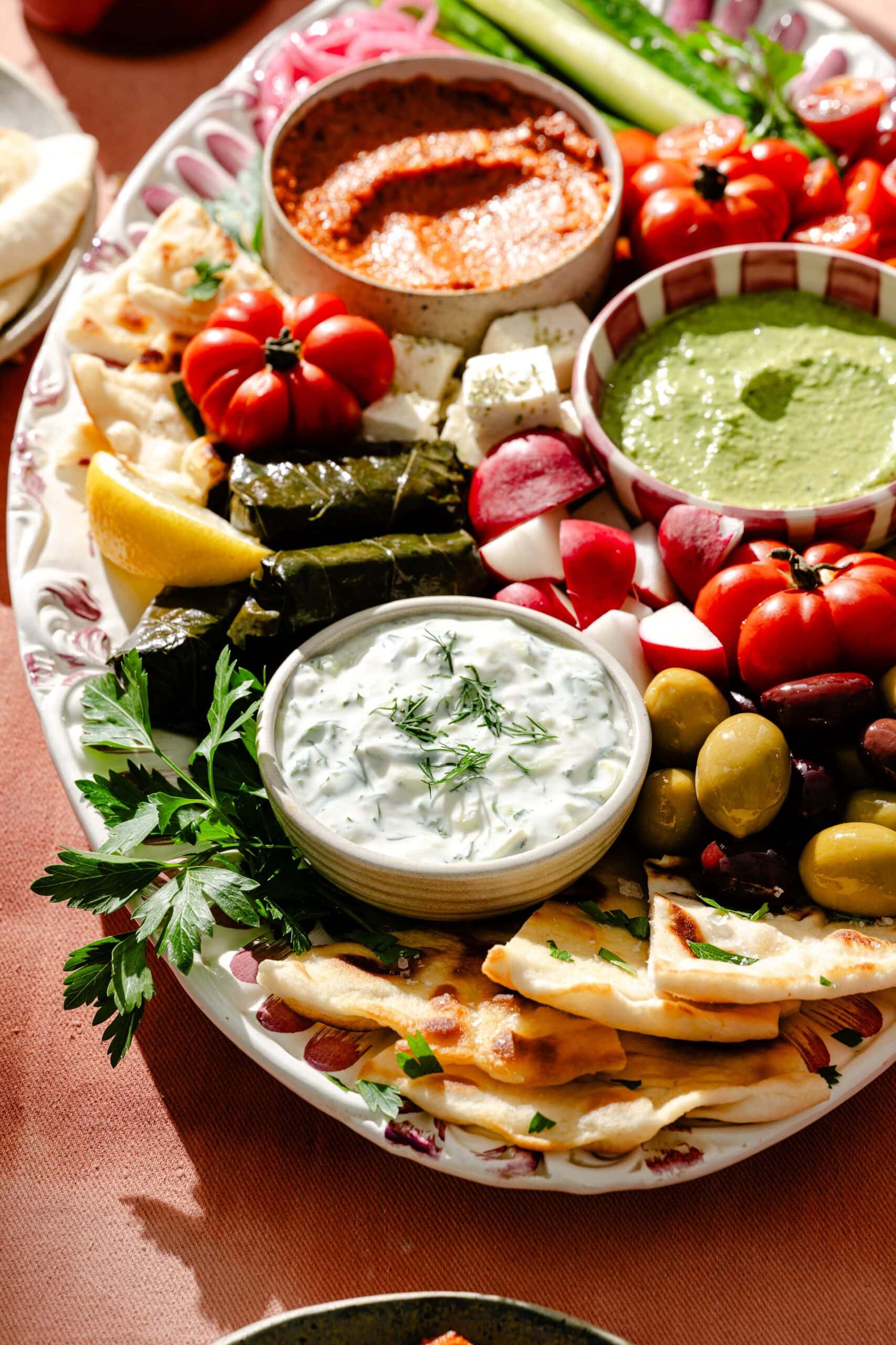 A platter with tzatziki, green sauce, red dip, olives, grape leaves, cherry tomatoes, radishes, sliced cucumbers, herbs, lemon, and pita bread, arranged decoratively on a patterned plate.