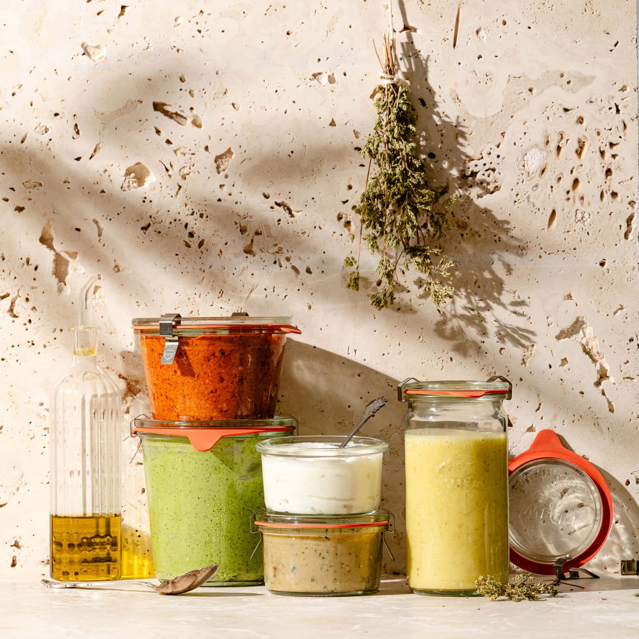 Jars of colorful sauces and a small oil bottle are arranged on a stone surface, with herbs hanging on the wall. Sunlight casts shadows, creating a rustic kitchen vibe.
