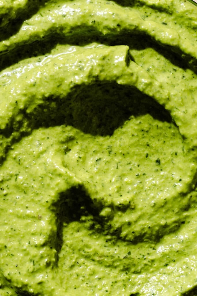Close-up of a thick, creamy green sauce or dip with visible herb flecks and swirled texture, possibly pesto or guacamole.