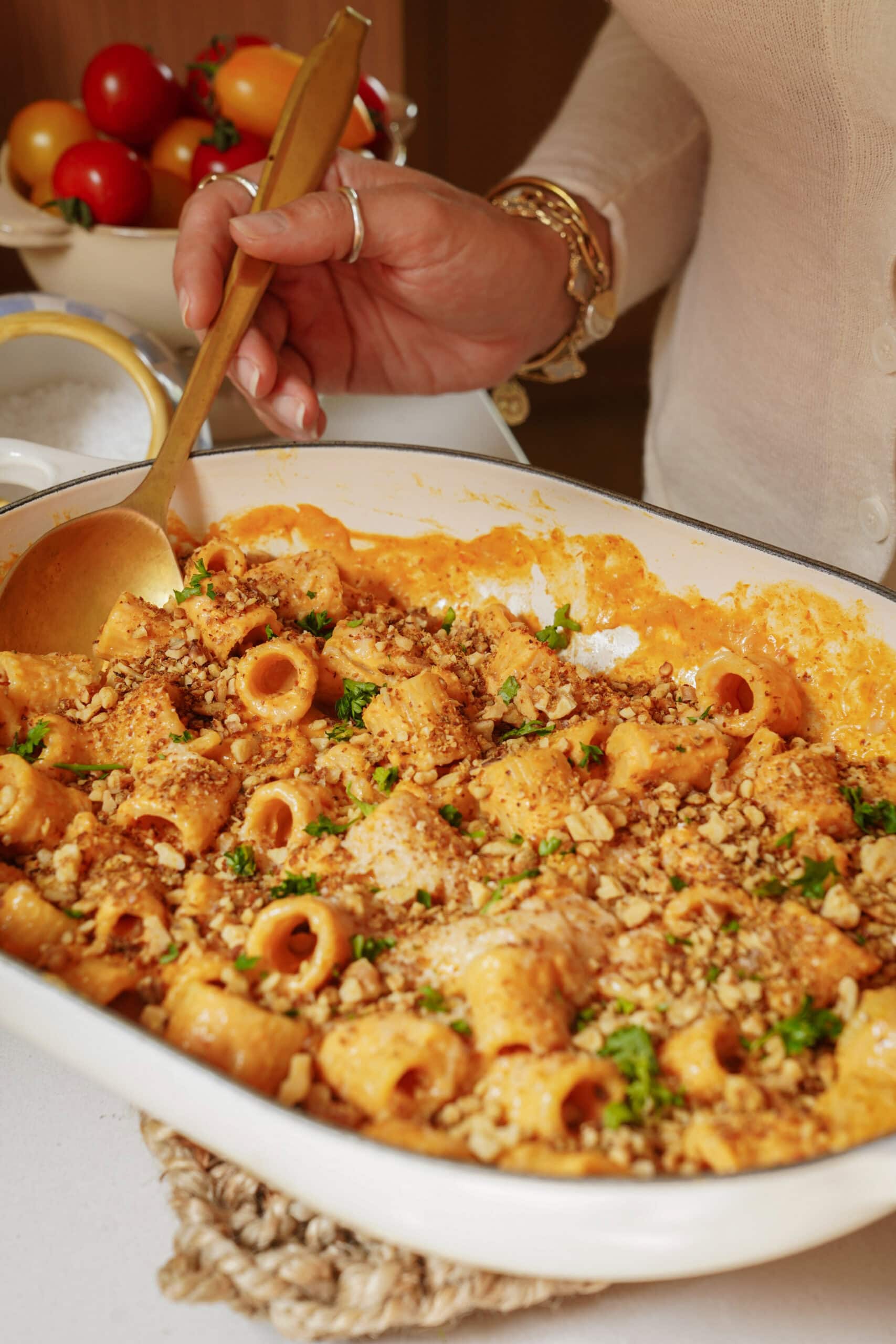 A person serves baked pasta with creamy sauce and breadcrumbs from a white dish using a gold spoon. Fresh tomatoes sit in a bowl in the background.