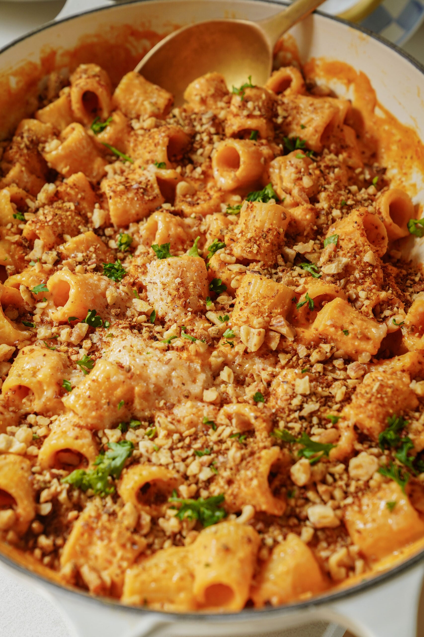 A close-up of creamy rigatoni pasta in a skillet, topped with breadcrumbs, chopped herbs, and a golden sauce, with a serving spoon resting inside.