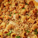 Rigatoni pasta in a creamy orange sauce, topped with chopped nuts, breadcrumbs, and fresh parsley. The dish appears baked and close-up, showing the texture and garnish clearly.