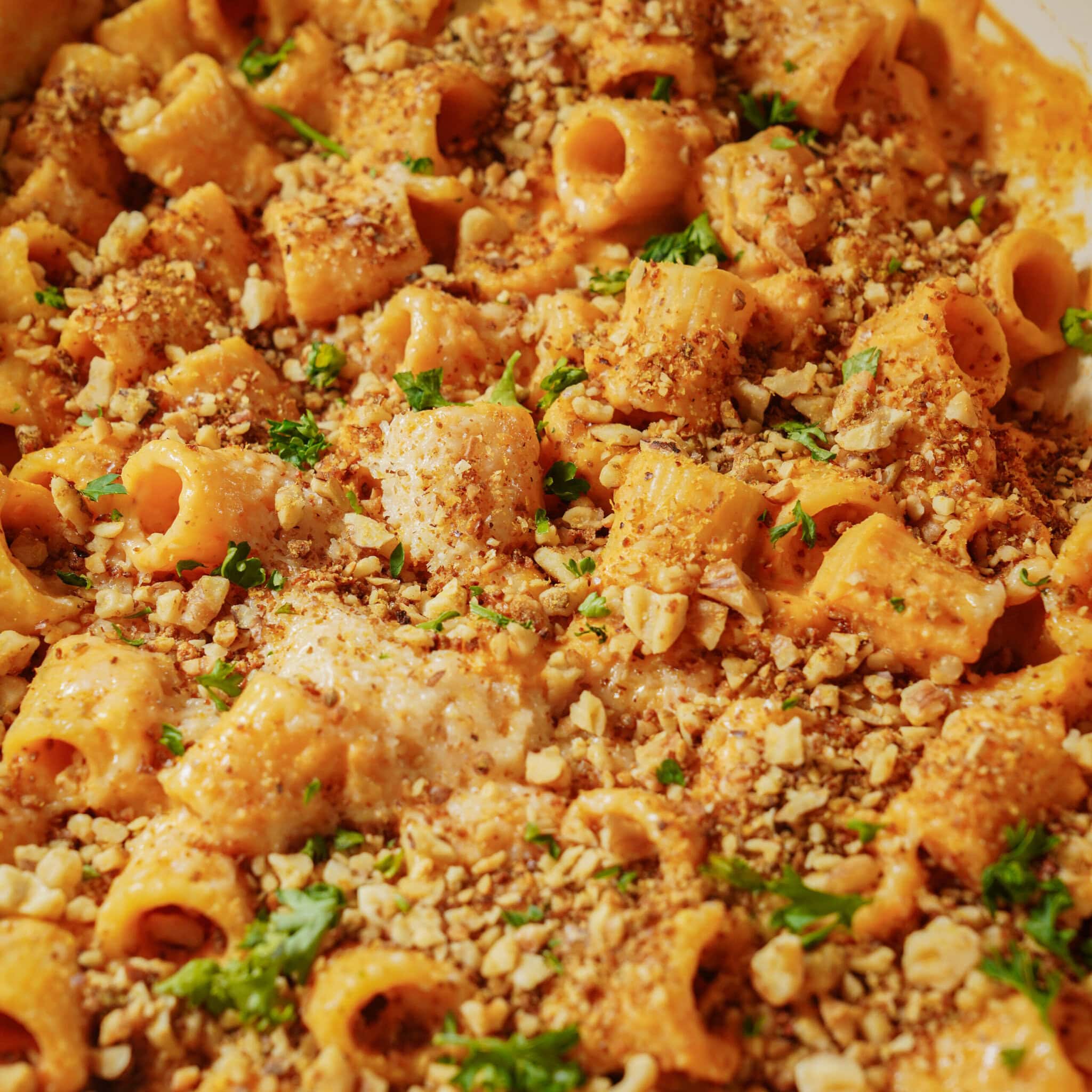 Rigatoni pasta in a creamy orange sauce, topped with chopped nuts, breadcrumbs, and fresh parsley. The dish appears baked and close-up, showing the texture and garnish clearly.