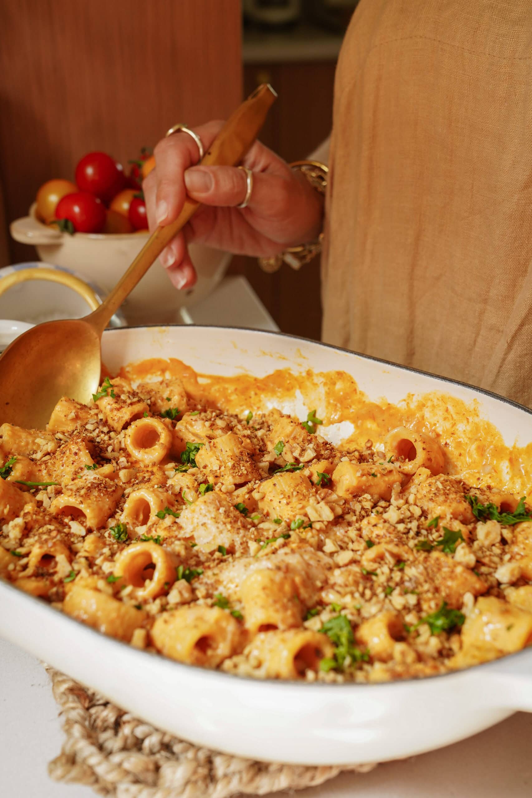 A person holds a gold spoon, serving creamy baked pasta topped with breadcrumbs and herbs from a white dish. Fresh tomatoes are in a bowl in the background.