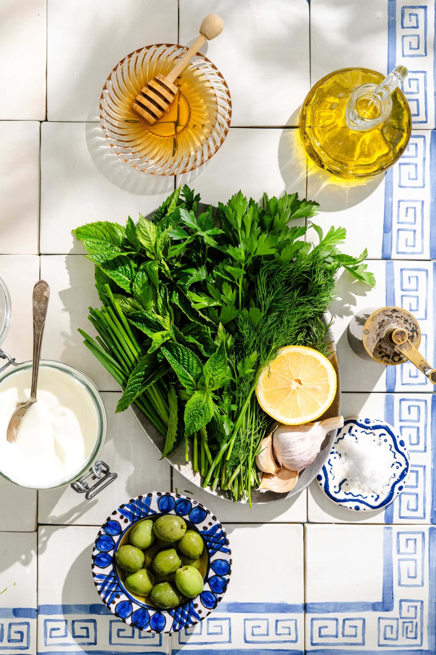 A plate of fresh herbs, half a lemon, garlic, salt, olives, honey, yogurt, mustard, and olive oil are arranged on a white tiled surface with a blue decorative border.