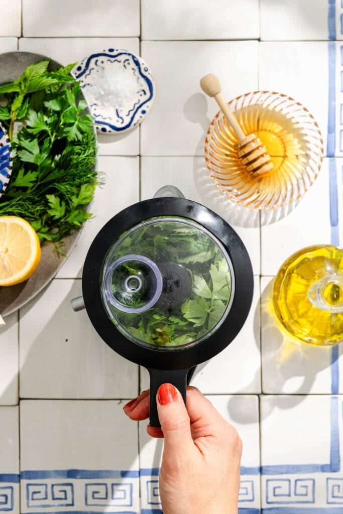A hand holds a small blender filled with fresh herbs. Nearby are a plate with herbs and lemon, a bowl of honey with a dipper, and a bottle of olive oil, all on a tiled countertop.