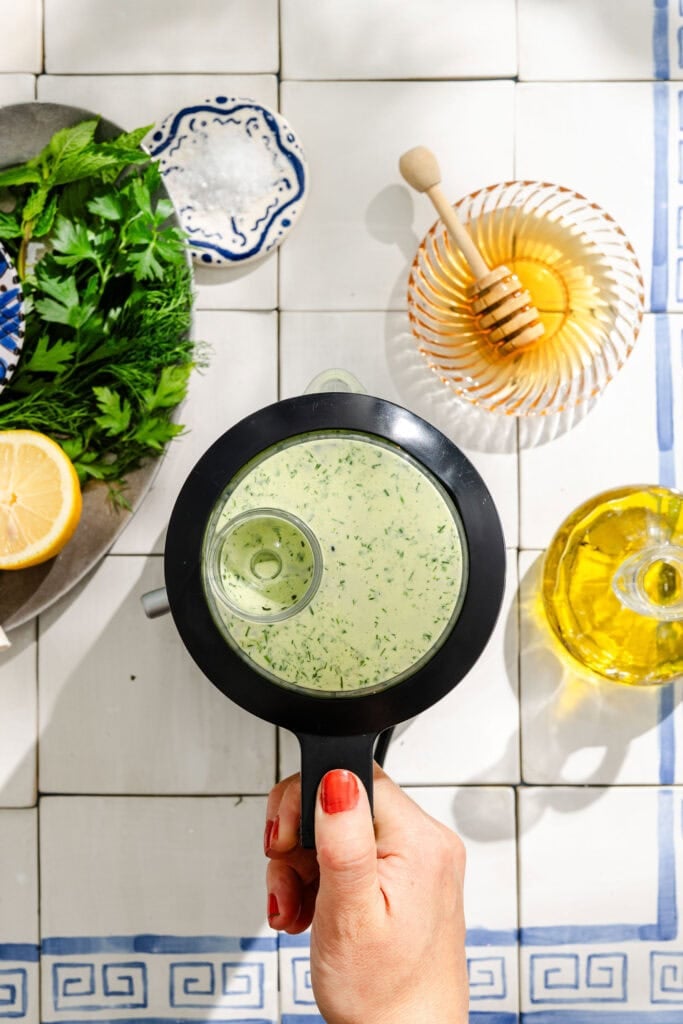 A hand holds a small blender filled with green herbed dressing above a tiled surface, surrounded by fresh herbs, lemon, olive oil, and honey.