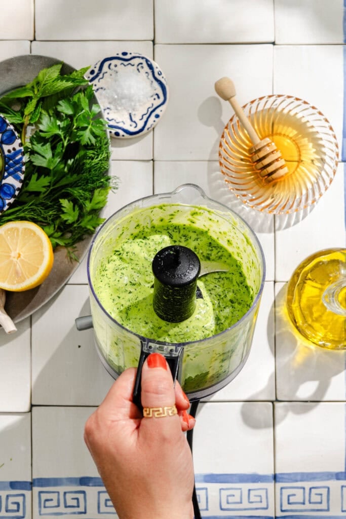 A hand holds a food processor with creamy green sauce inside, surrounded by fresh herbs, half a lemon, honey dipper in a bowl, and a bottle of olive oil on a tiled countertop.