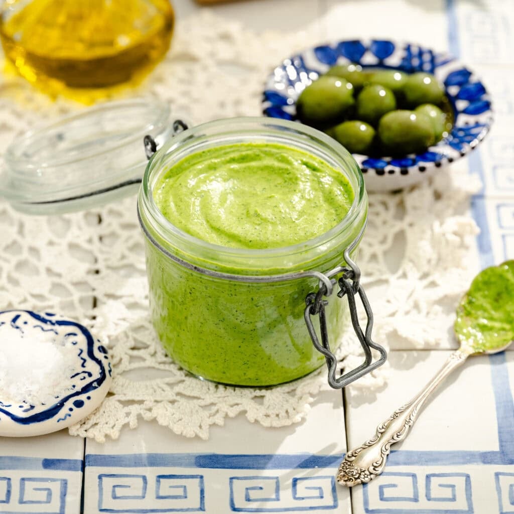 A glass jar of green sauce on a lace doily, with a spoon of sauce beside it. Nearby are a plate of green olives, a dish of salt, and a bottle of olive oil on a blue and white tiled table.