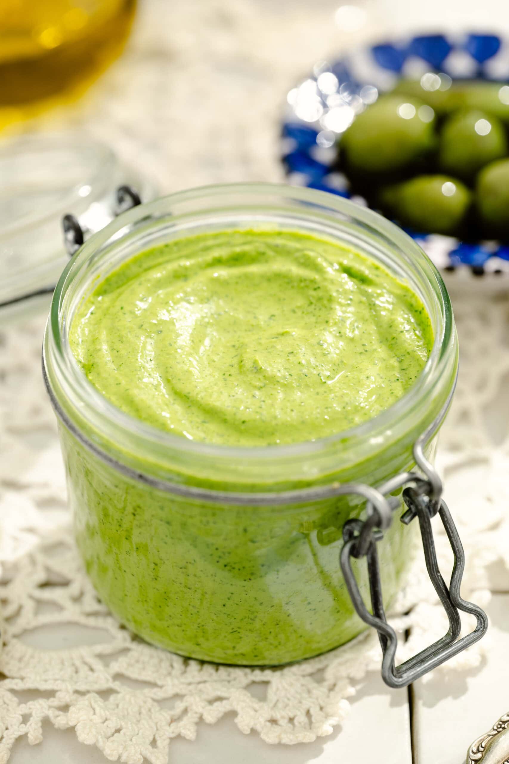 A glass jar filled with creamy green sauce sits on a lace tablecloth, with green olives and a partial blue patterned dish visible in the background.
