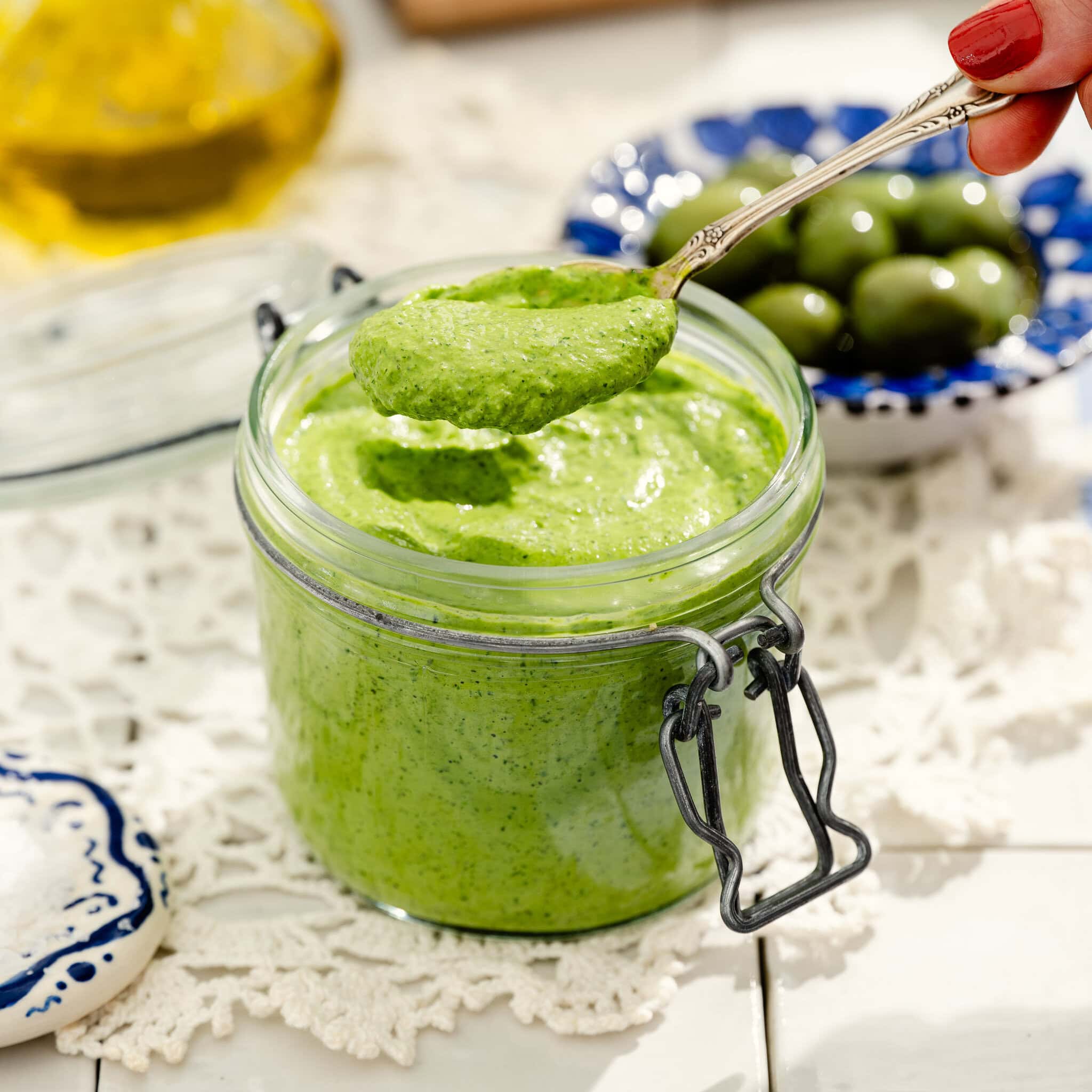 A hand holds a spoonful of vibrant green pesto above an open glass jar. In the background are a bowl of green olives, a white lace cloth, and part of a bottle of olive oil.