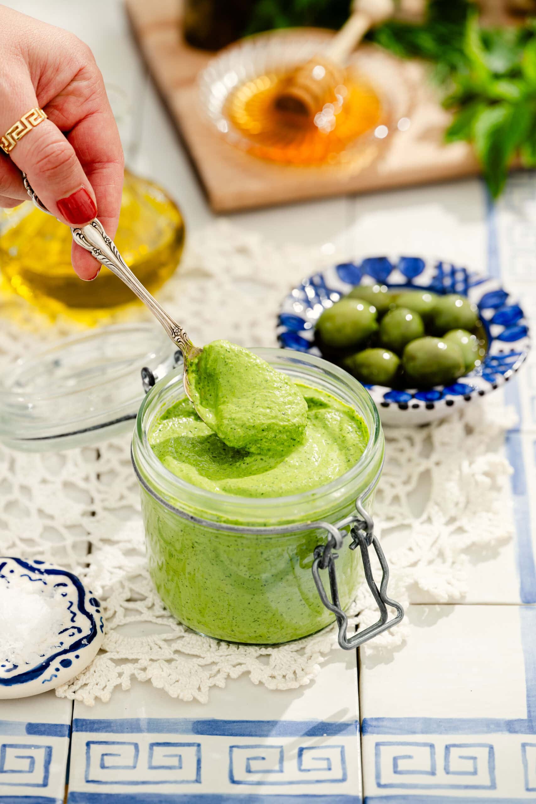A hand holding a spoon dips into a jar of green sauce. Nearby are green olives, a dish of salt, olive oil, and honey on a table with blue and white tiles and a lace doily.