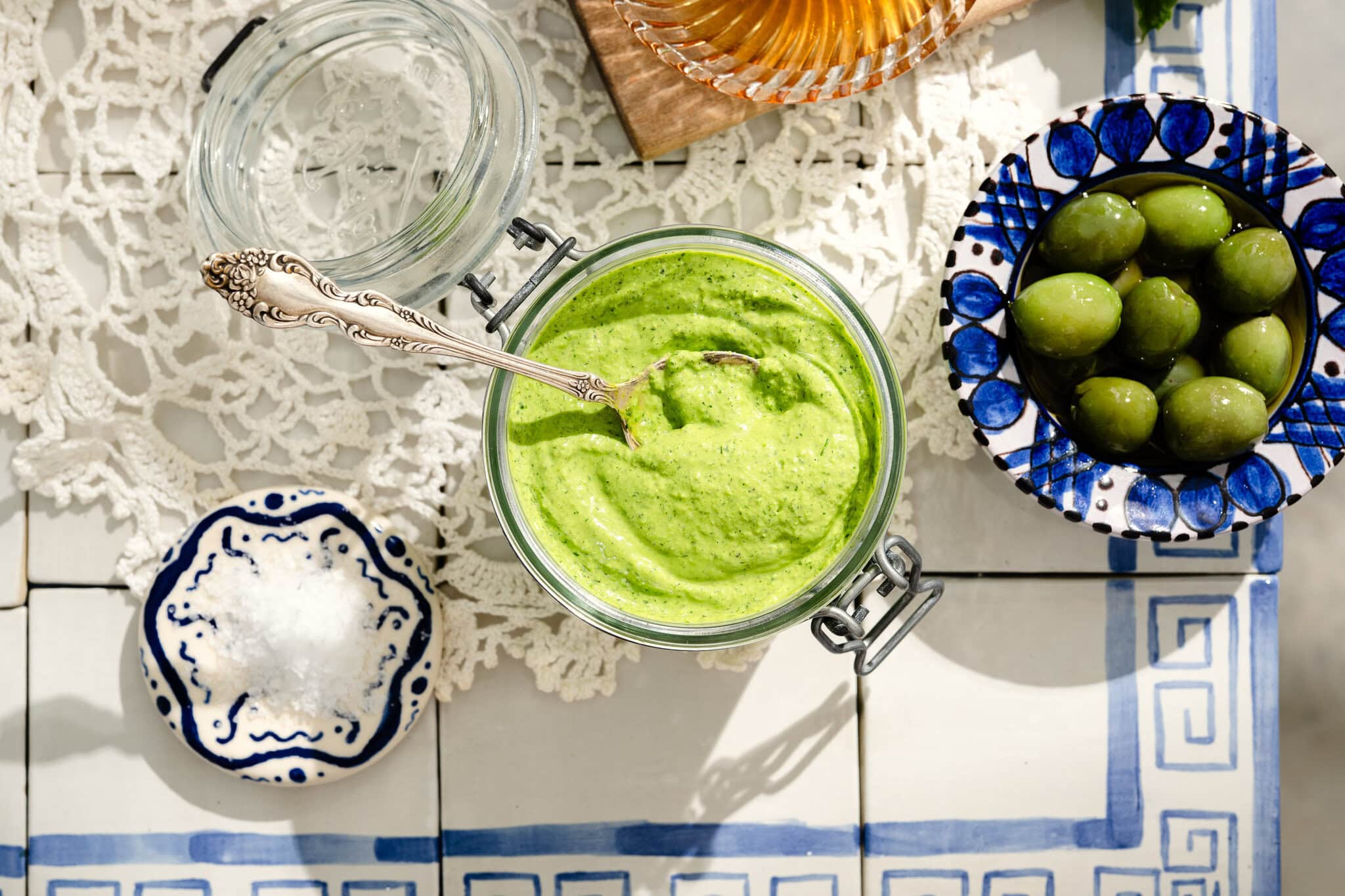 A jar of green sauce with a spoon sits on a tiled table with a bowl of green olives, a dish of salt, and a glass of water. The table is partially covered with a crochet lace doily.