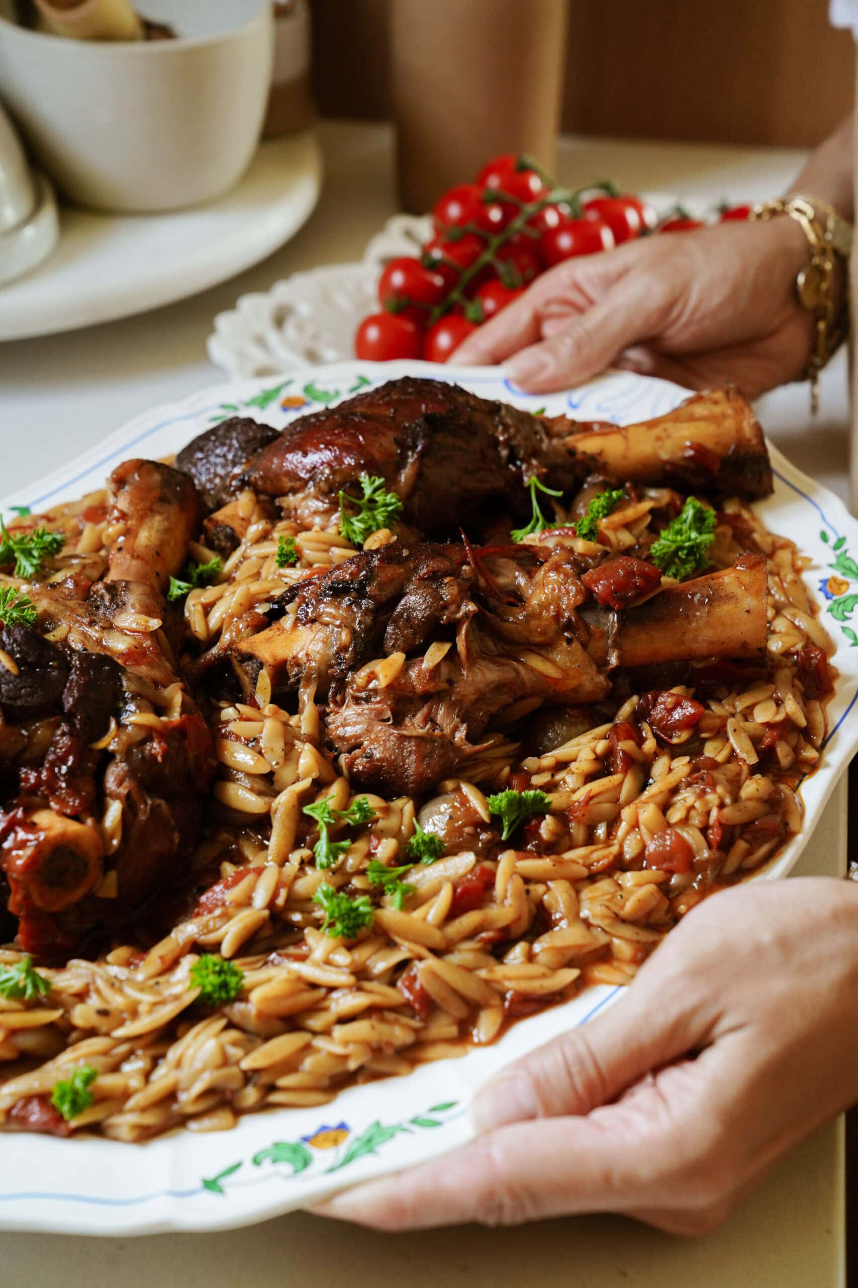 A person holds a platter of braised chicken drumsticks served over orzo pasta with tomato sauce, garnished with fresh parsley. In the background, another person holds a plate with a bunch of cherry tomatoes.