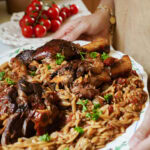 A person holds a platter of braised lamb shanks served over orzo pasta with tomato sauce and fresh parsley. A bunch of tomatoes sits in the background.