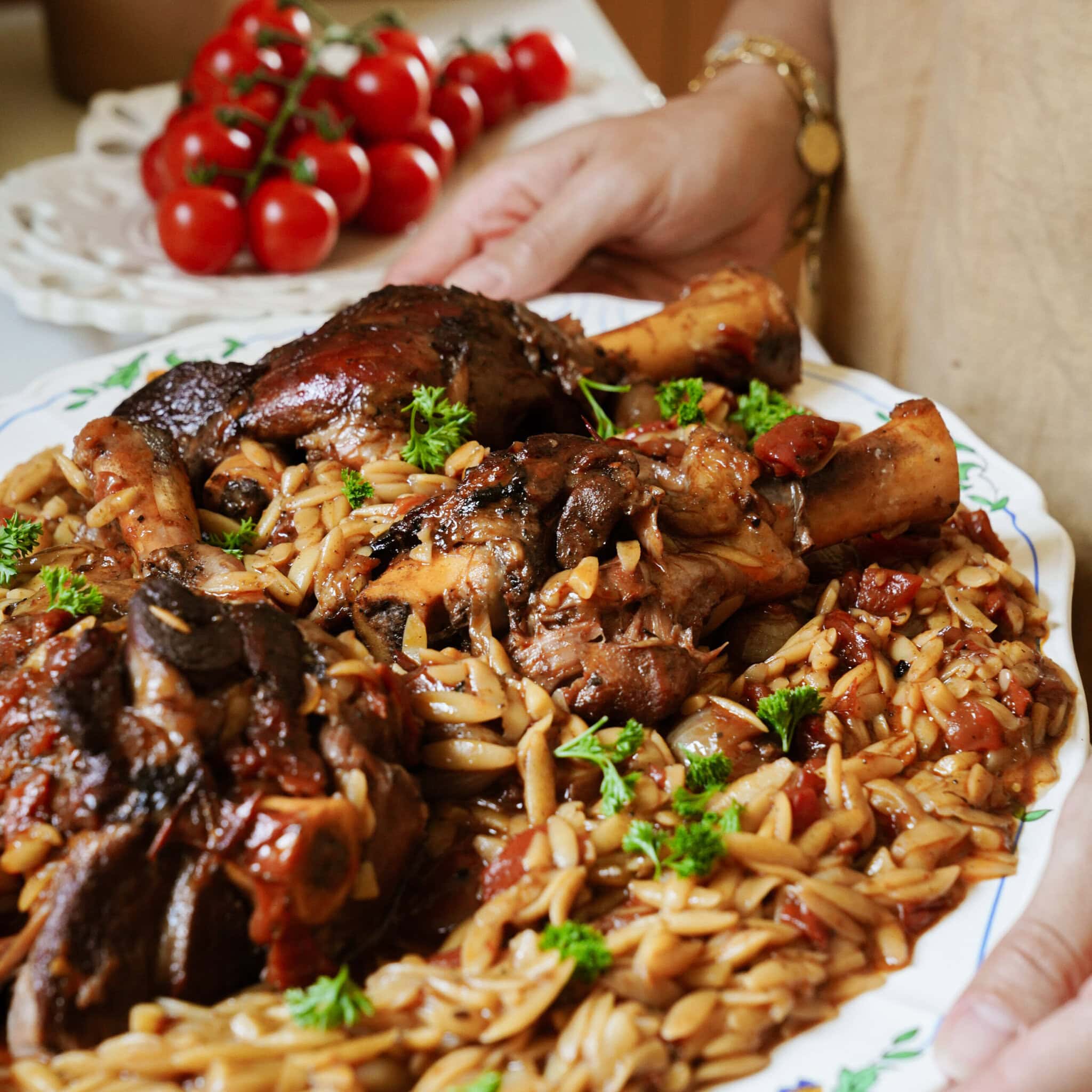 A person holds a platter of braised lamb shanks served over orzo pasta with tomato sauce and fresh parsley. A bunch of tomatoes sits in the background.