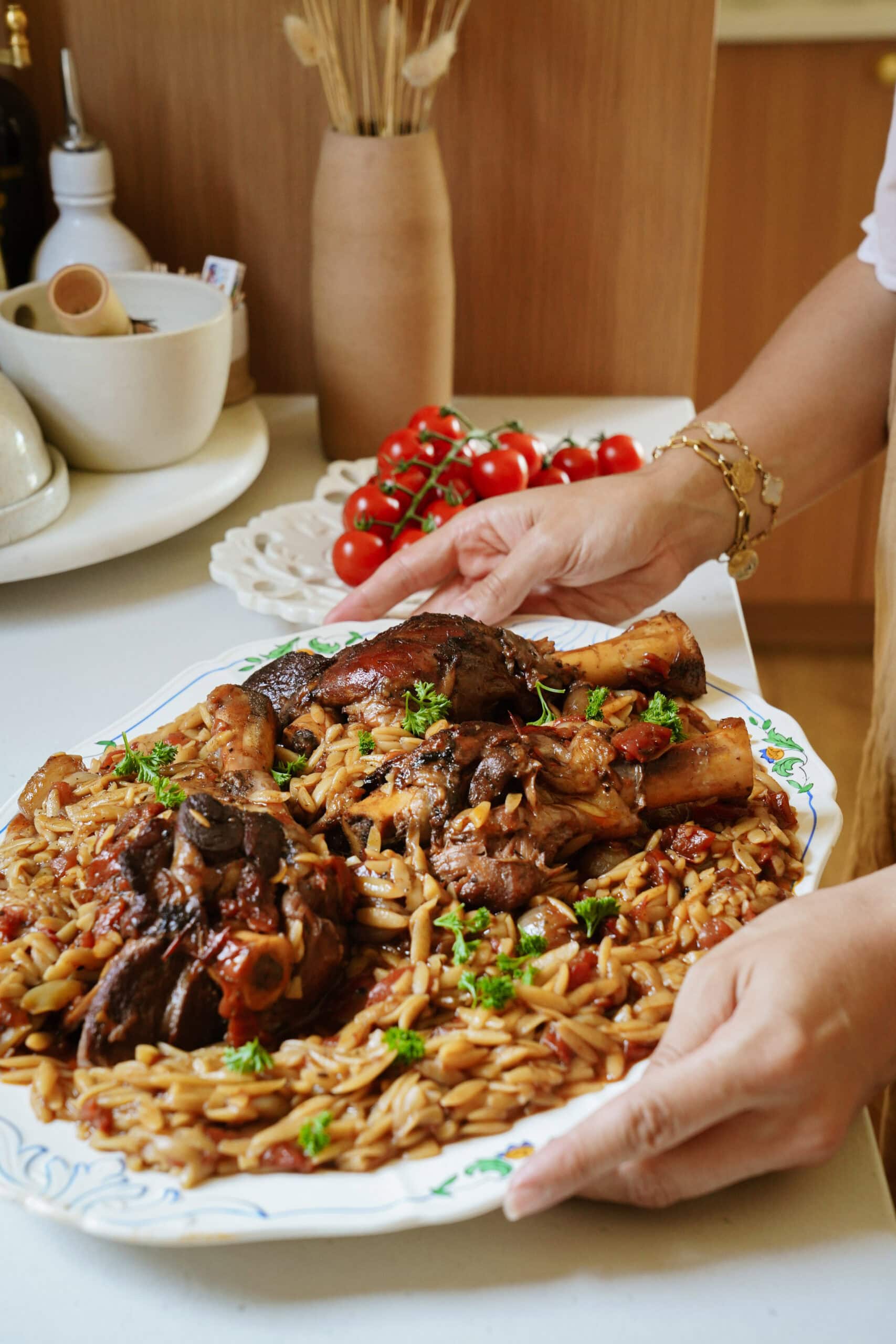 A person holds a platter of braised meat and orzo pasta garnished with parsley, with fresh tomatoes and kitchen items visible in the background.