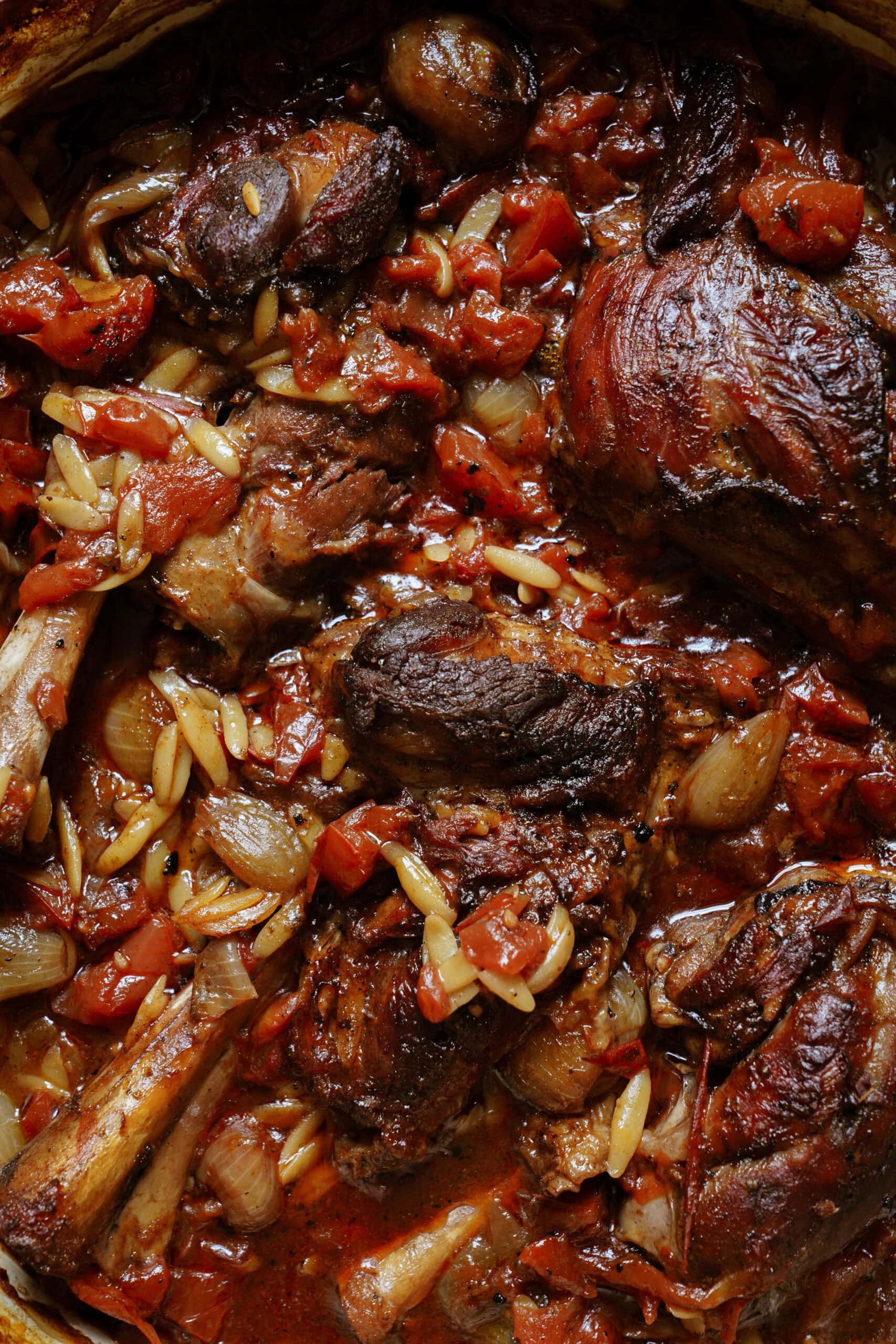 Braised lamb shanks in a rich tomato sauce with whole shallots, slivered almonds, and visible pasta or orzo, all cooked together until tender and flavorful.