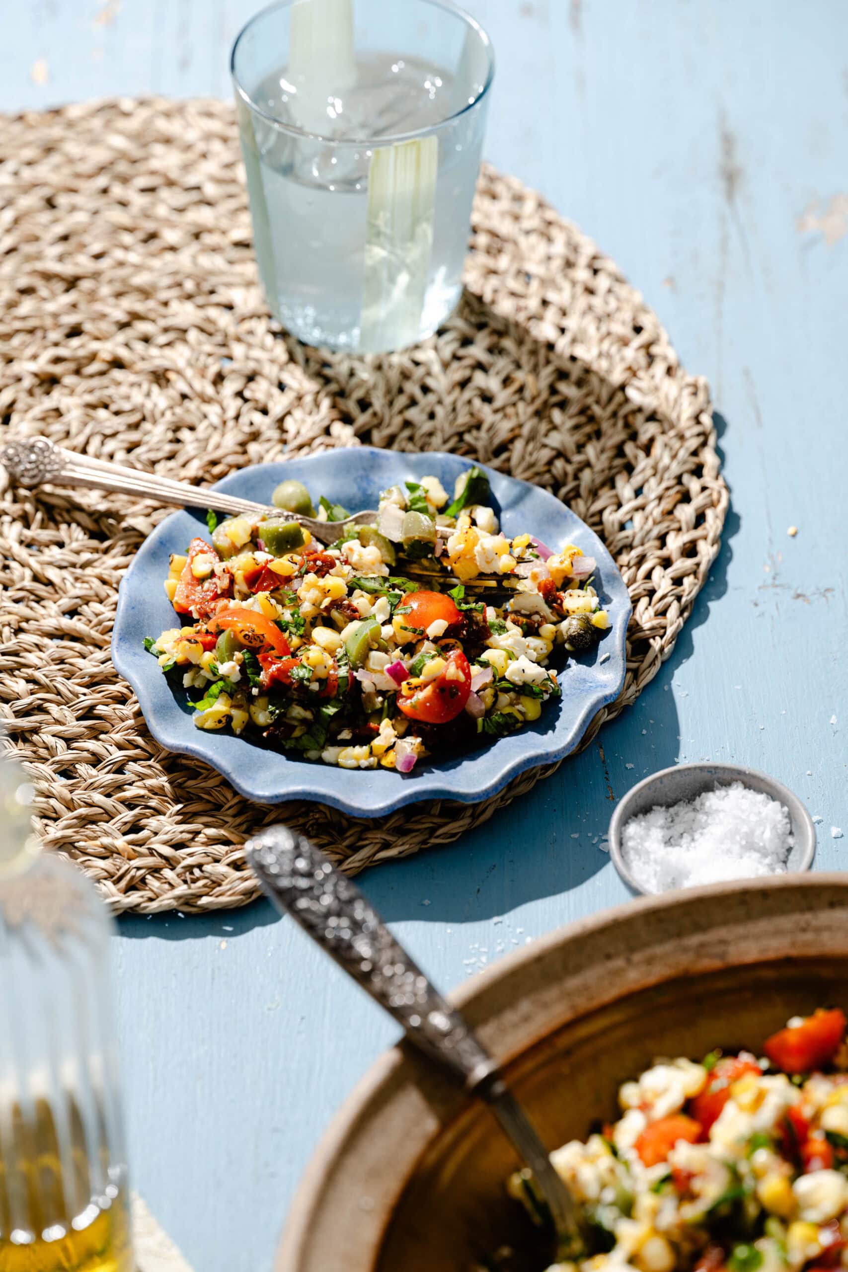 A small plate of colorful vegetable salad on a woven placemat, with a glass of water and a bowl of salt nearby on a blue table under bright natural light.