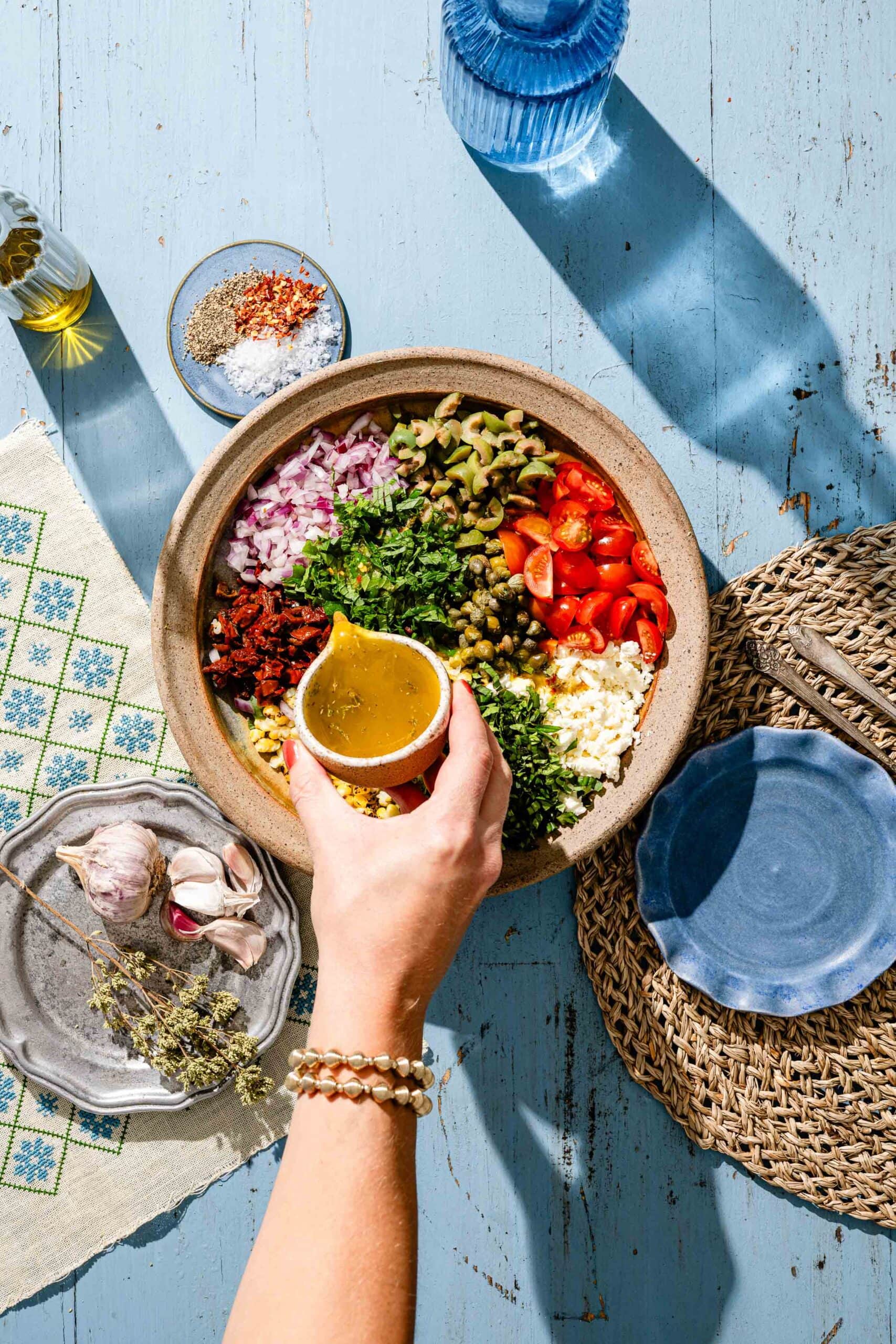 A hand pours dressing over a bowl filled with chopped veggies, herbs, and cheese on a blue table, surrounded by garlic, seasonings, plates, and a patterned cloth.