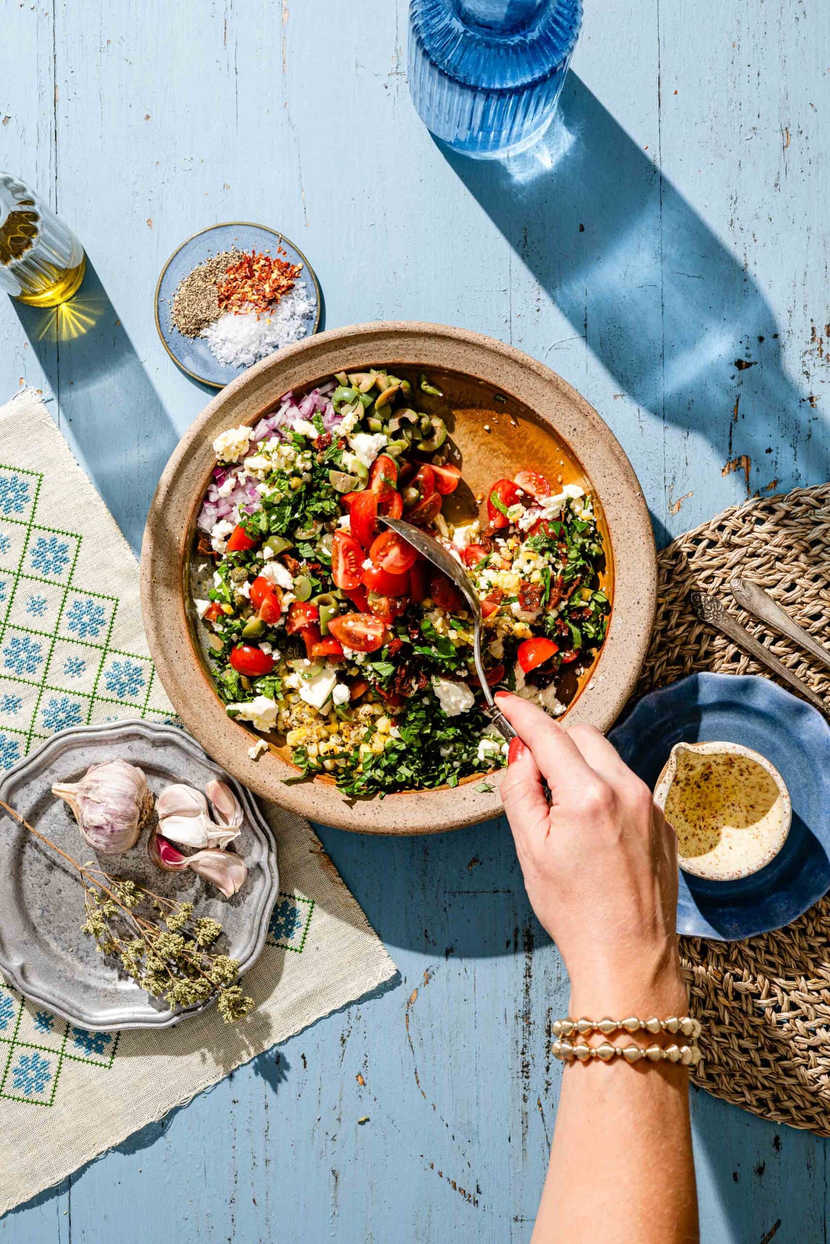 A hand mixes a colorful salad with tomatoes, herbs, and cheese in a bowl on a blue wooden table, surrounded by garlic, oil, spices, and a beverage.